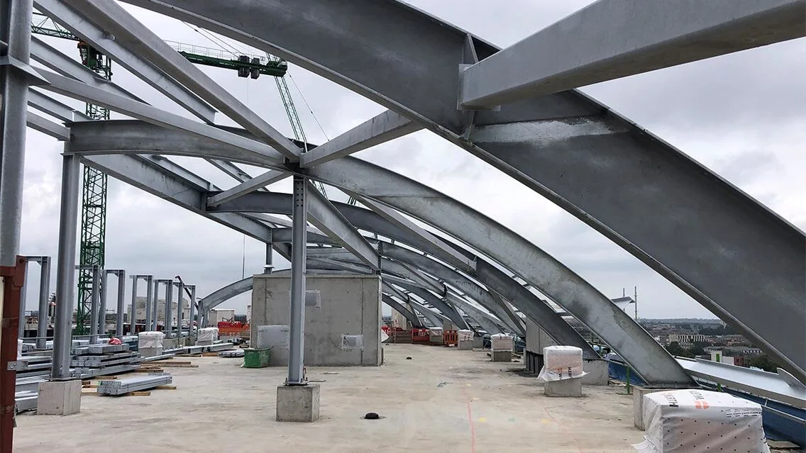 Close-up of a building's internal steel structure during construction, with arched beams and support columns visible against a cloudy sky.