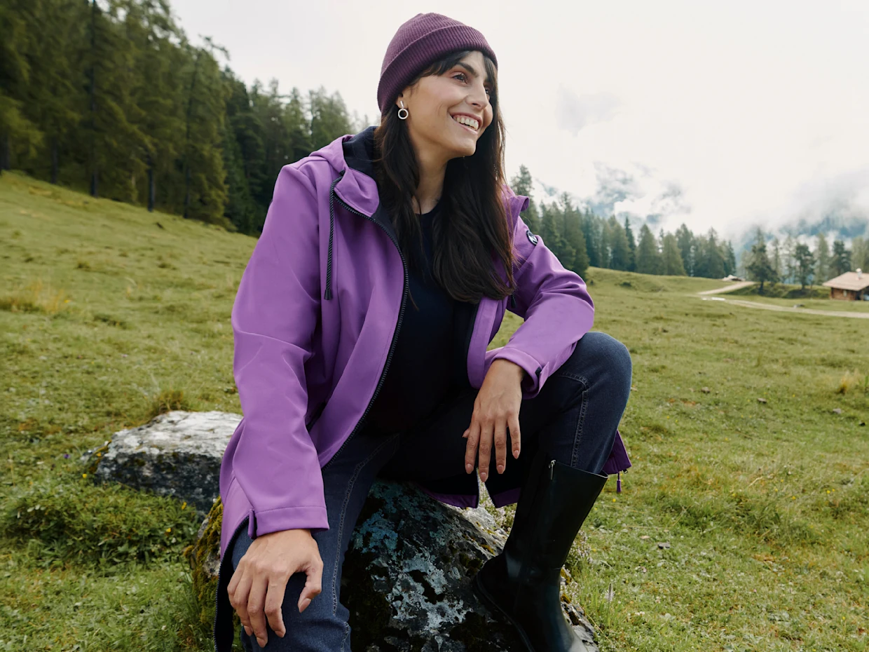 Ein Model sitzt auf einem Stein in einer grünen Wiese, trägt eine lila Softshelljacke, Mütze, Jeans und schwarze Stiefel. Ein Model sitzt auf einem Stein in einer grünen Wiese, trägt eine lila Softshelljacke, Mütze, Jeans und schwarze Stiefel.