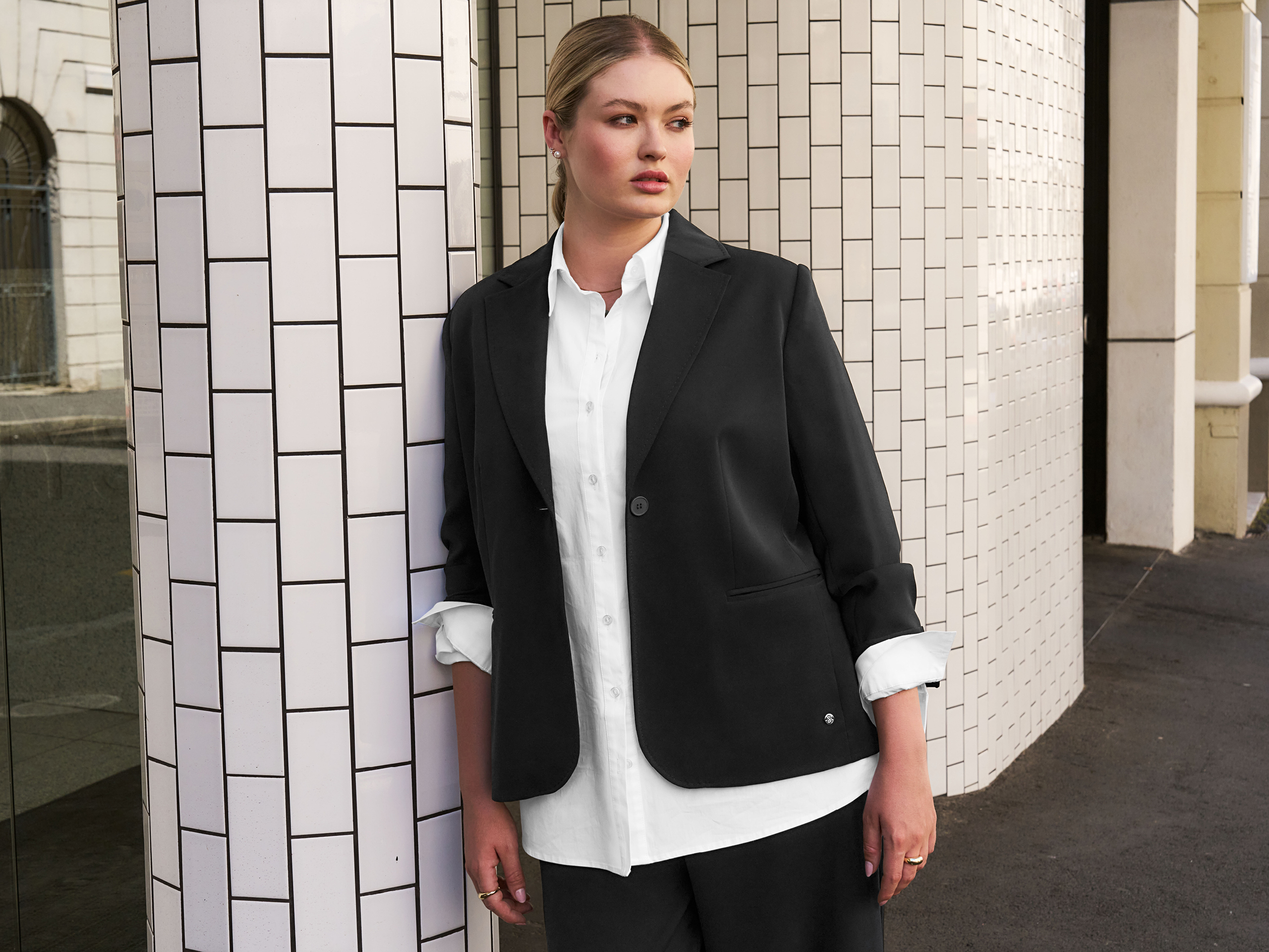 Ein Model steht an einer Wand mit weißen Kacheln, trägt einen schwarzen Blazer und ein weißes Hemd.