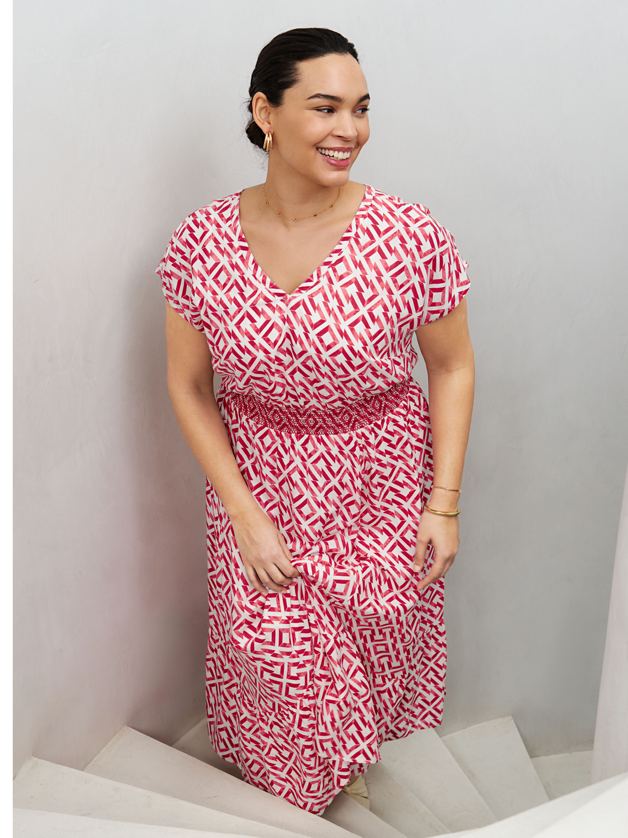 Ein Model trägt ein Kleid mit einemrot-weißen, geometrischen Muster und steht auf einer Treppe.
Das Kleid hat kurze Ärmel und einen V-Ausschnitt. Die Taille ist mit einem elastischen Band betont.