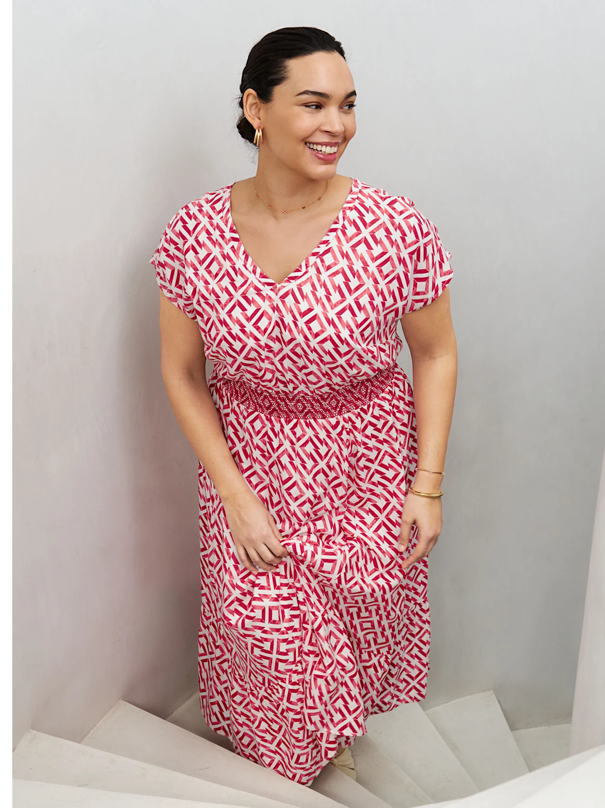 Ein Model trägt ein Kleid mit einemrot-weißen, geometrischen Muster und steht auf einer Treppe.
Das Kleid hat kurze Ärmel und einen V-Ausschnitt. Die Taille ist mit einem elastischen Band betont.