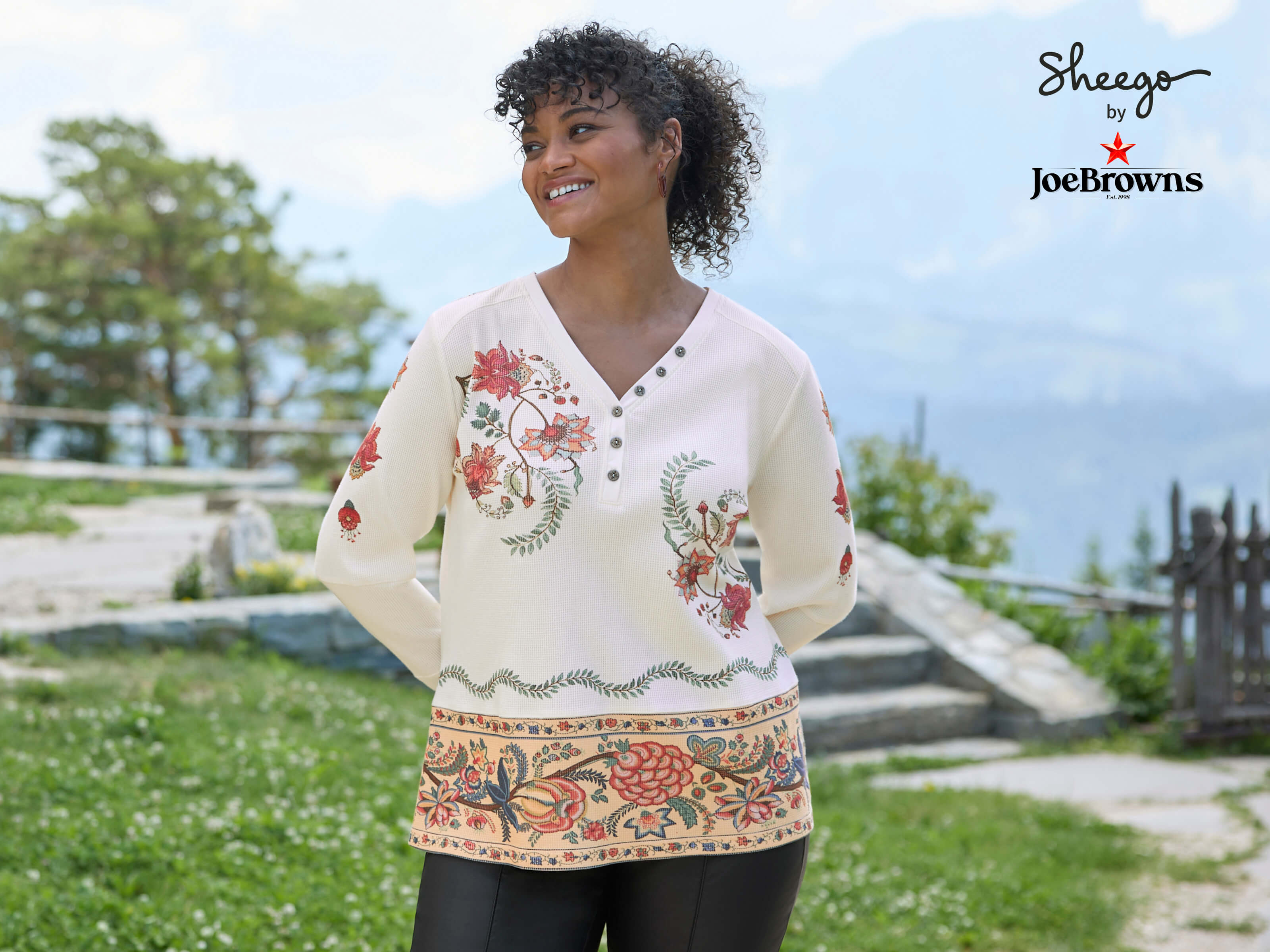 Ein Model trägt ein champagner-rostrot-bedrucktes Langarmshirt mit floralem Muster und eine schwarze Hose. 