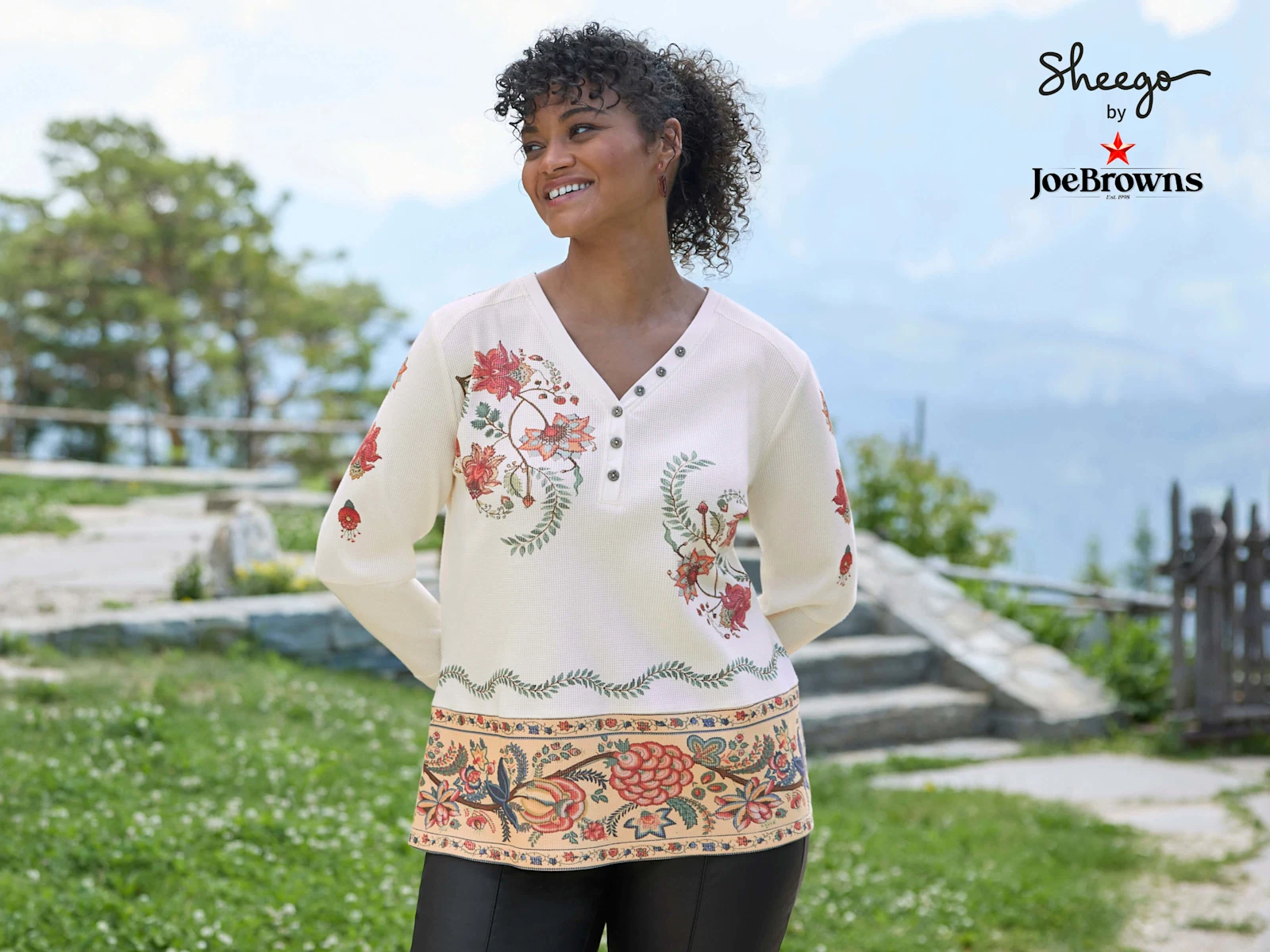 Ein Model trägt ein champagner-rostrot-bedrucktes Langarmshirt mit floralem Muster und eine schwarze Hose.