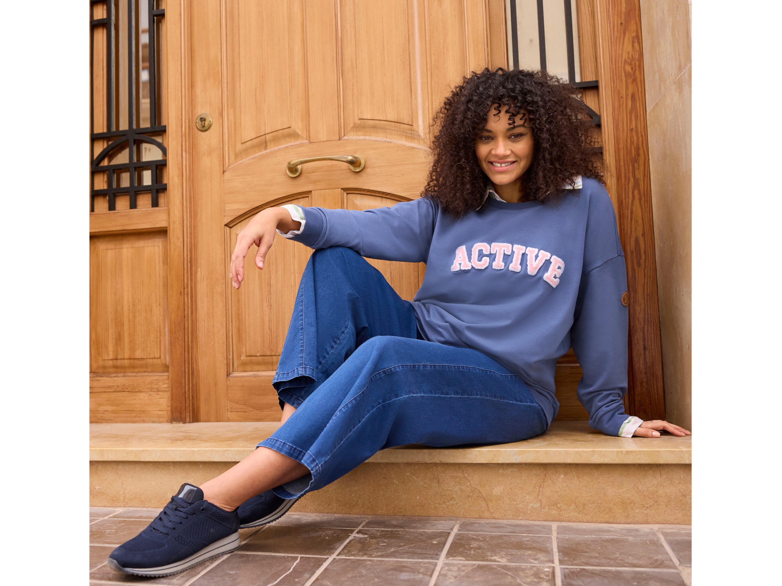 Ein Model sitzt vor einer Tür, sie trägt ein rauchblaues Sweatshirt mit Print über einer gestreiften Bluse und eine weite Jeans in blue-stone-washed
