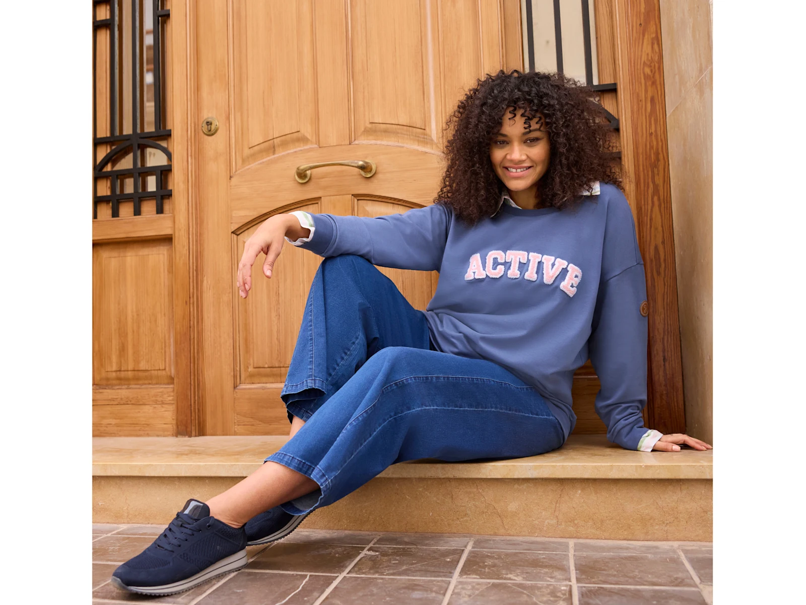 Ein Model sitzt vor einer Tür, sie trägt ein rauchblaues Sweatshirt mit Print über einer gestreiften Bluse und eine weite Jeans in blue-stone-washed