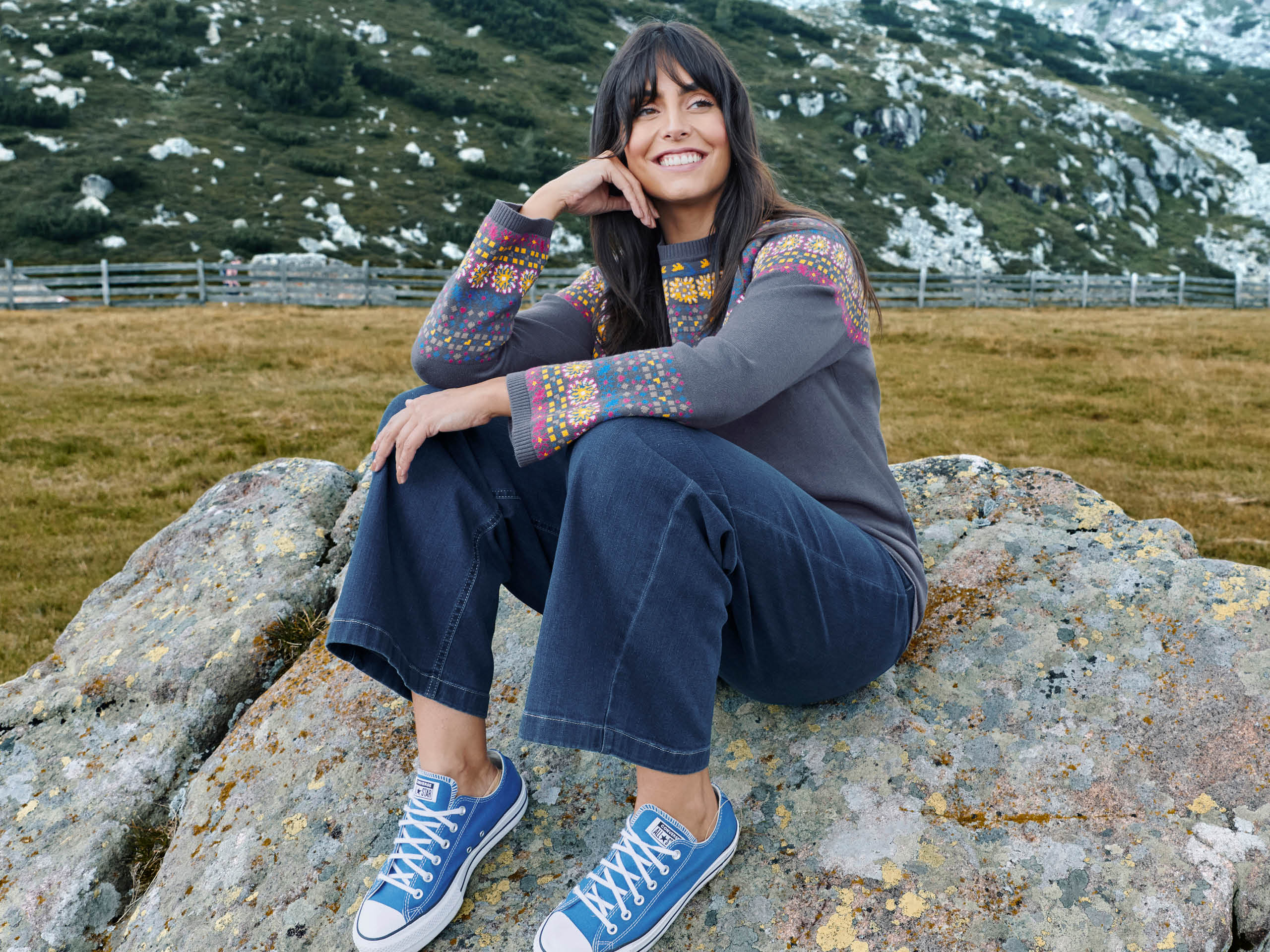 Ein Model sitzt in einem bergigen Gelände mit Wiese auf einem Stein. Sie trägt einen rauchblauen Pullover mit Norweger-Jacquard-Muster, eine blaue Bootscut Jeans und blaue Sneaker.