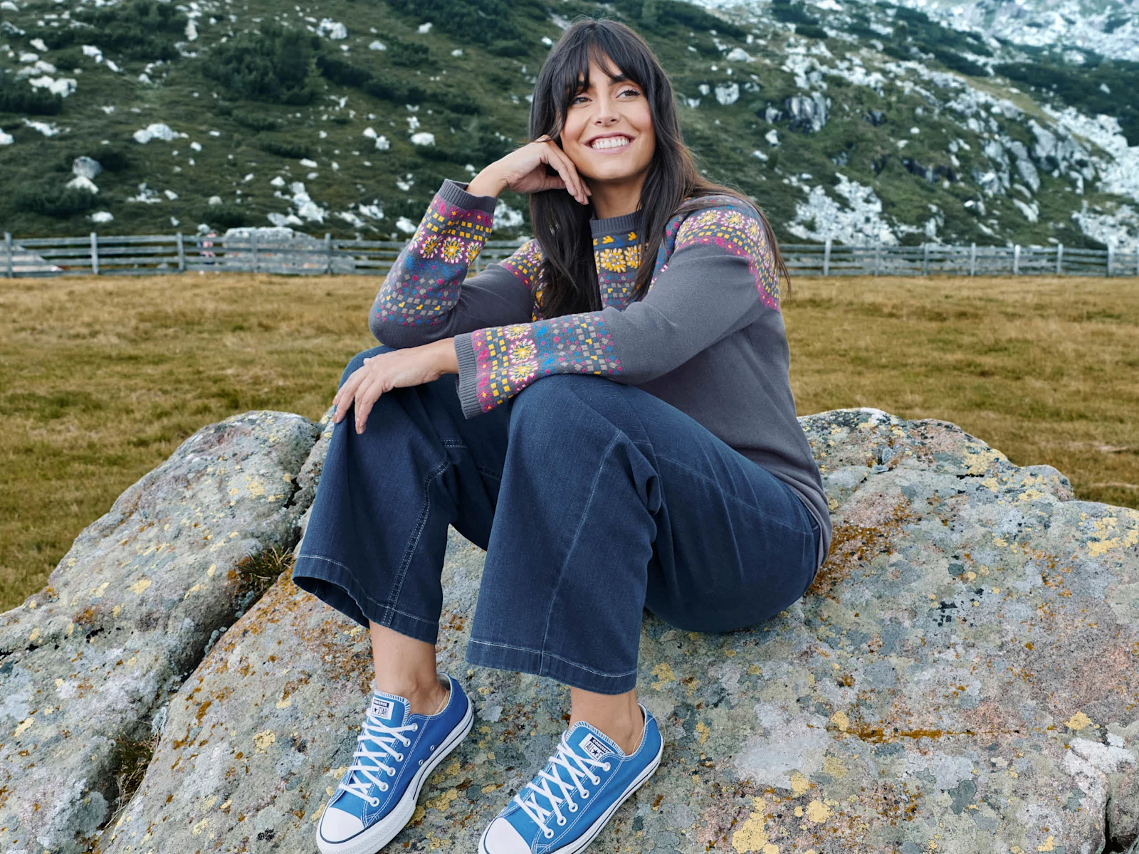 Ein Model sitzt in einem bergigen Gelände mit Wiese auf einem Stein. Sie trägt einen rauchblauen Pullover mit Norweger-Jacquard-Muster, eine blaue Bootscut Jeans und blaue Sneaker.
