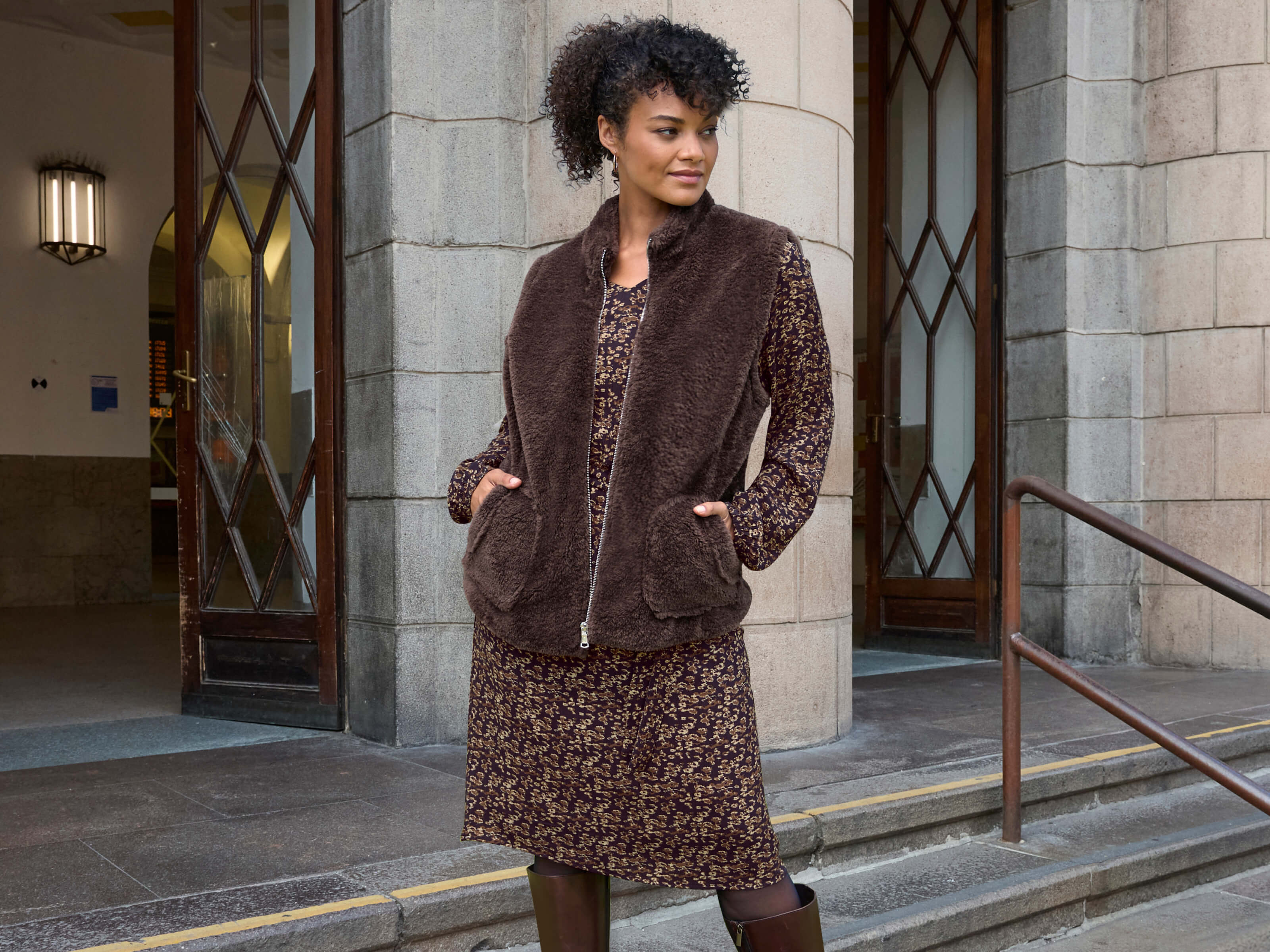 Ein Model trägt ein schoko-camel-bedrucktes Webkleid mit Minimalprint und eine braune Teddyweste mit Reißverschluss