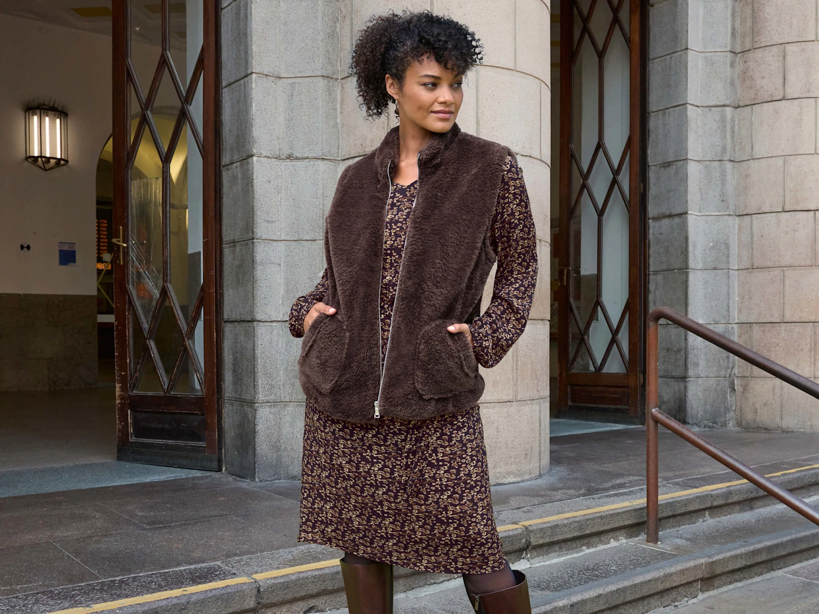 Ein Model trägt ein schoko-camel-bedrucktes Webkleid mit Minimalprint und eine braune Teddyweste mit Reißverschluss