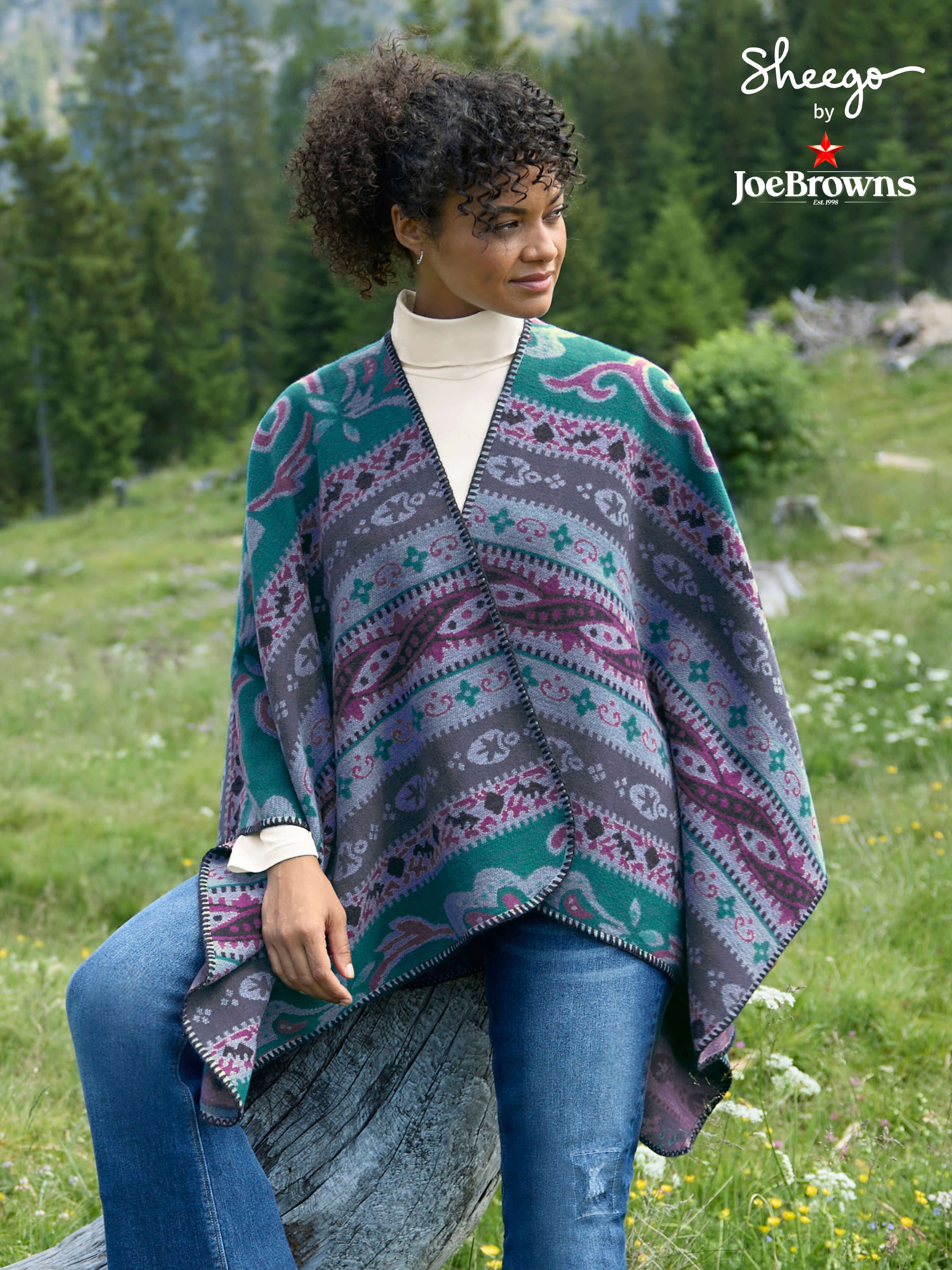 Ein Model trägt einen petrol-malve-gemustertn Poncho, ein champagnerfarbenes Rollkragenshirt und eine dark blue denim Bootcut-Jeans. Daneben ist das Sheego by Joe Browns Logo zu sehen und folgender Text: Warm ums Herz. Neues für den Winter