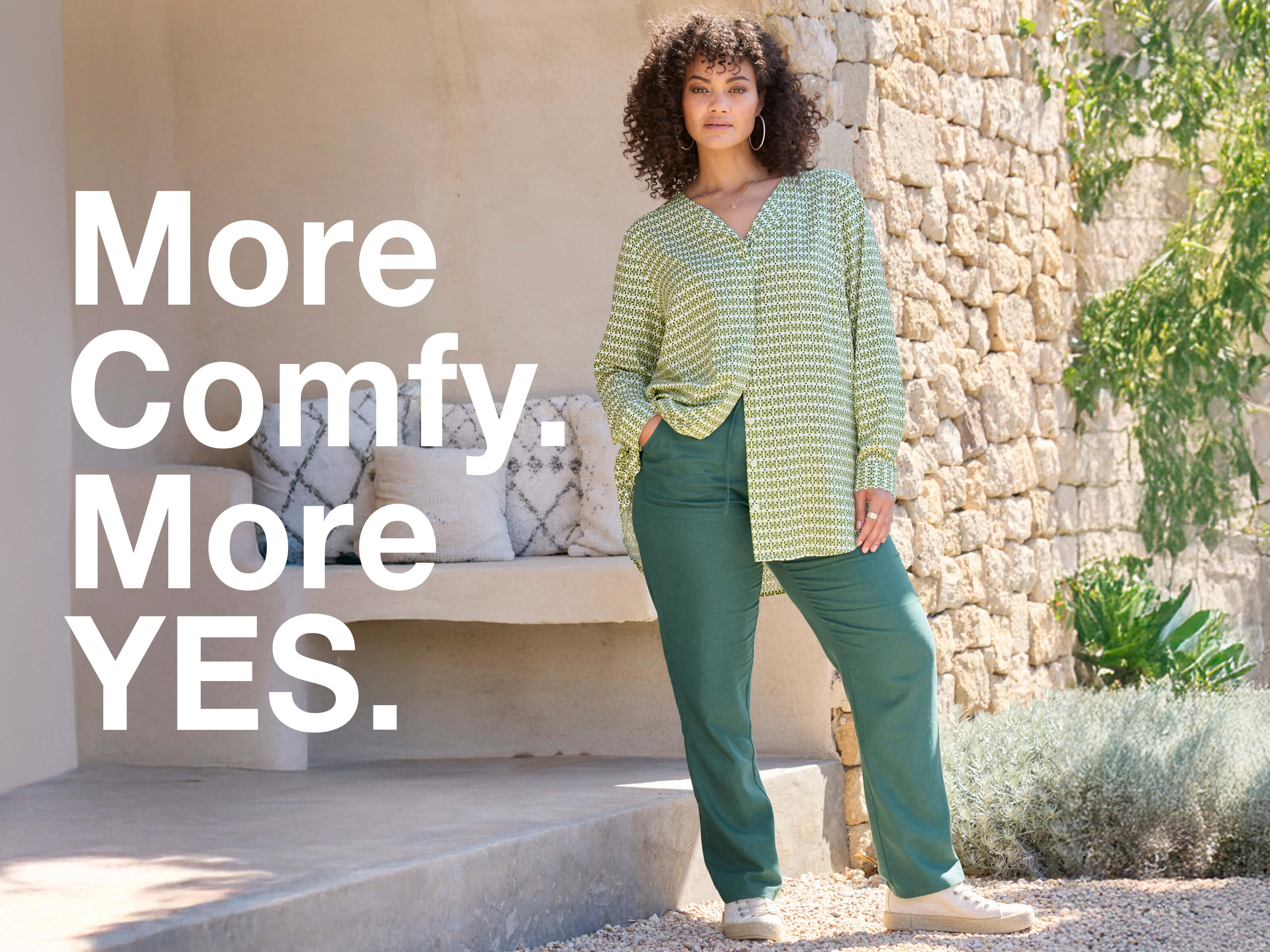 Ein Model in frühlingshafter Outdoor-Umgebung trägt eine  mossgrüne Jogg-Pants aus Leinen-Viskose-Mix und eine farblich passende Bluse.  Daneben folgender Text: More comfy, more yes.
