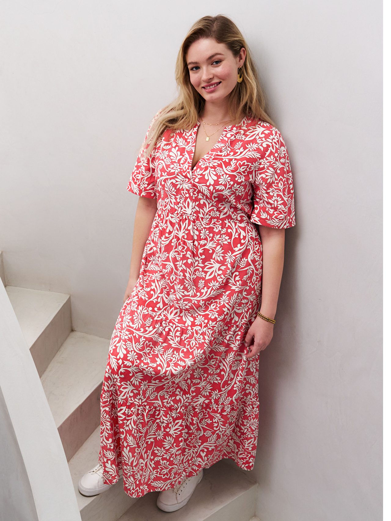 Ein Model steht auf einer Treppe und trägt ein weiß-rotes Maxikleid mit floralem Muster. Das Kleid hat kurze Ärmel und einen V-Ausschnitt.