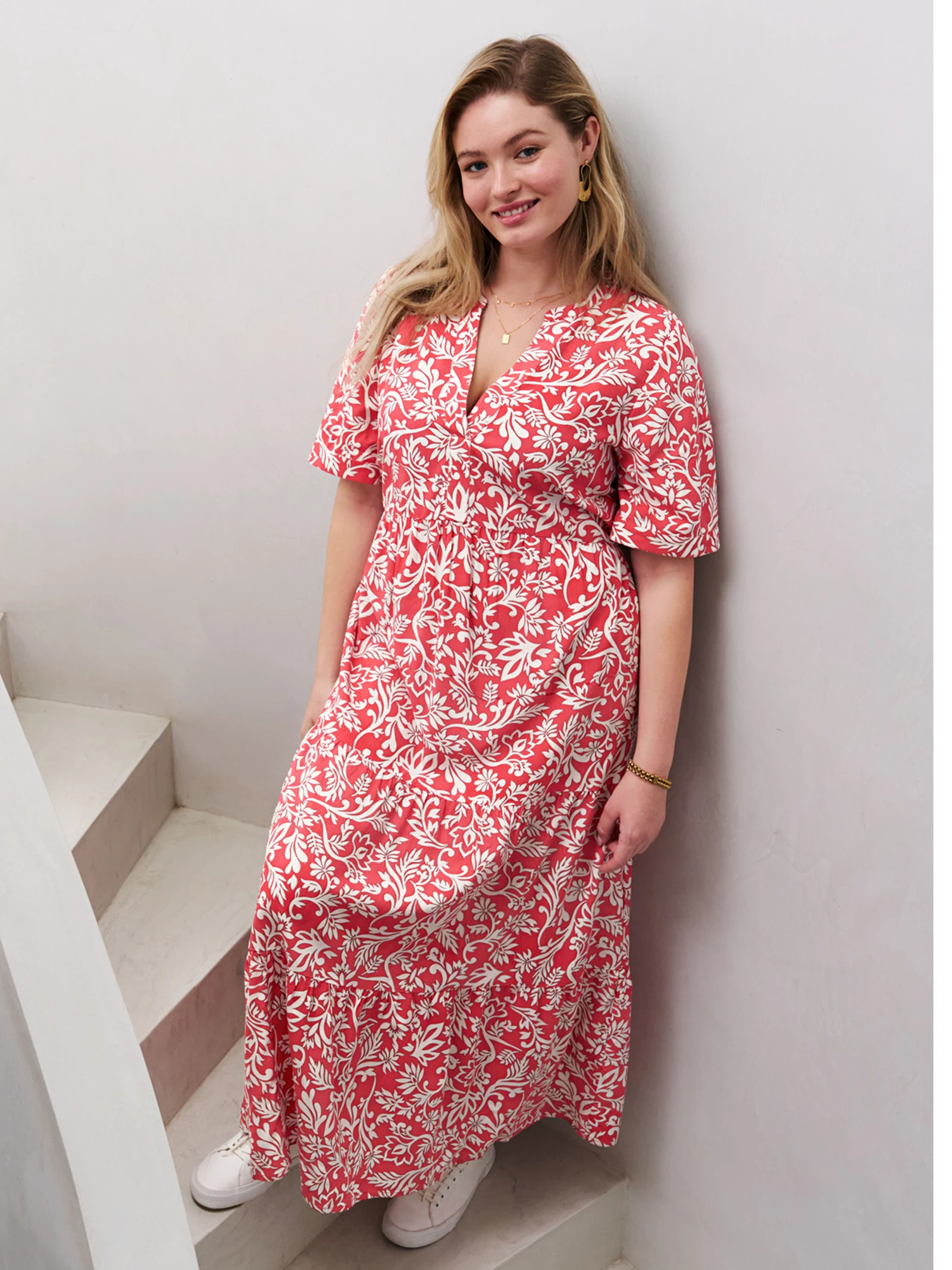 Ein Model steht auf einer Treppe und trägt ein weiß-rotes Maxikleid mit floralem Muster. Das Kleid hat kurze Ärmel und einen V-Ausschnitt.