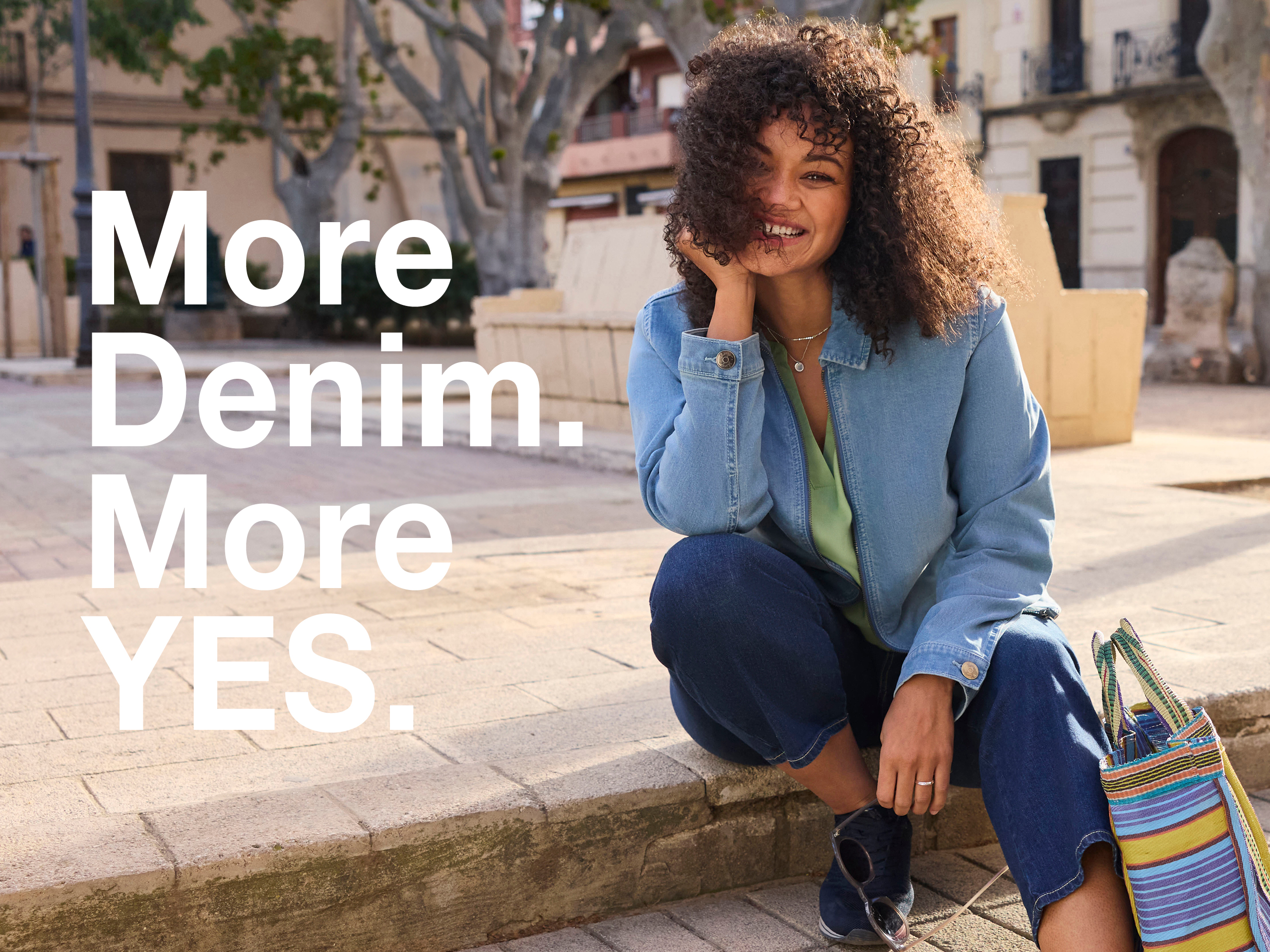 Ein Model in einer frühlingshaften City-Atmosphäre, sie trägt eine eucalyptus-fabene Bluse, eine verkürzt-geschnittenene Jeans in dark blue und eine blue bleached Jeansjacke. Daneben der Text: More Denim, more YES.