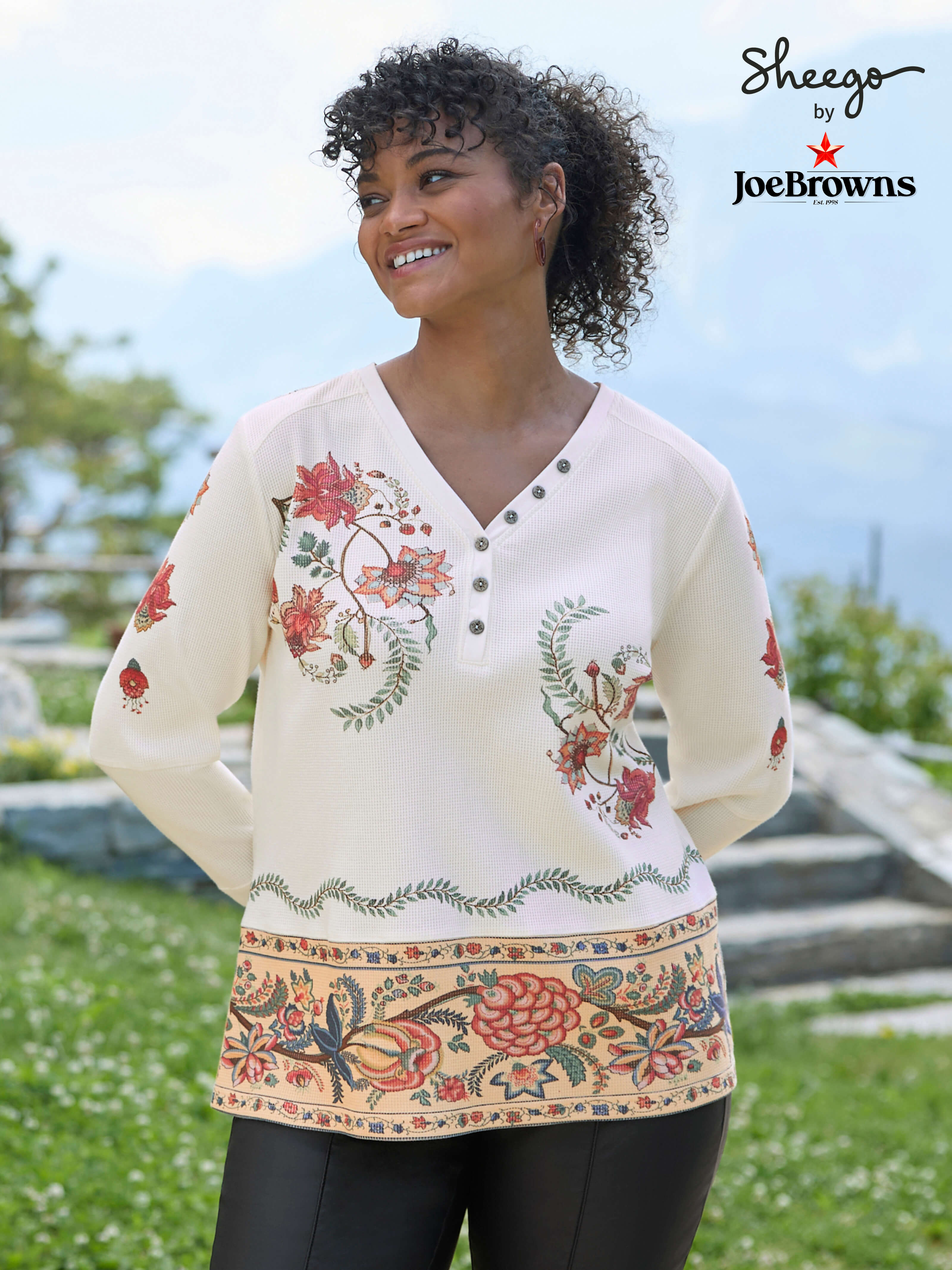 Ein Model trägt ein champagner-rostrot-bedrucktes Langarmshirt mit floralem Muster und eine schwarze Hose. 