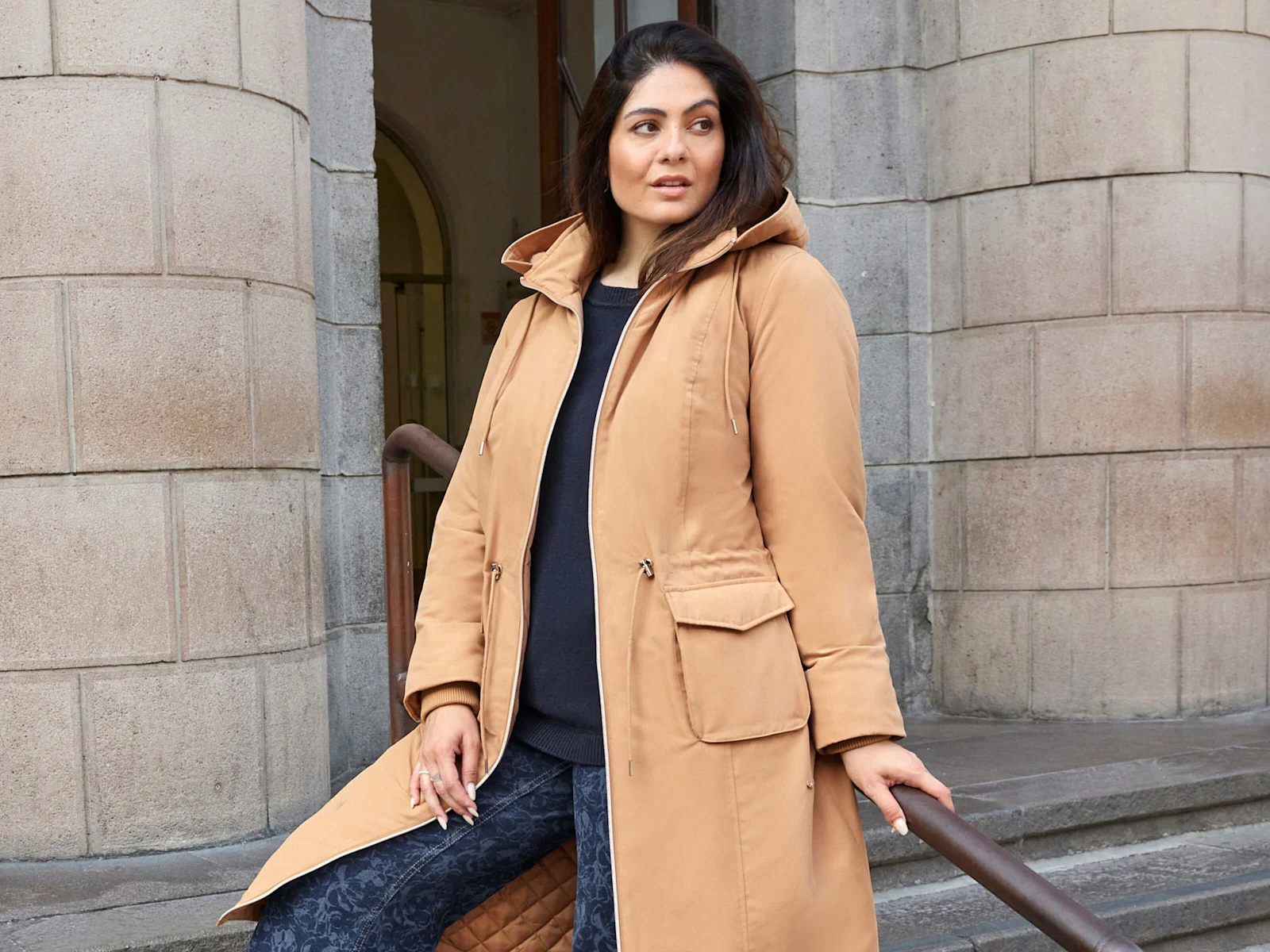 Ein Model trägt einen camel farbenen Parka in wärmender Qualität und einen blauen Pullover