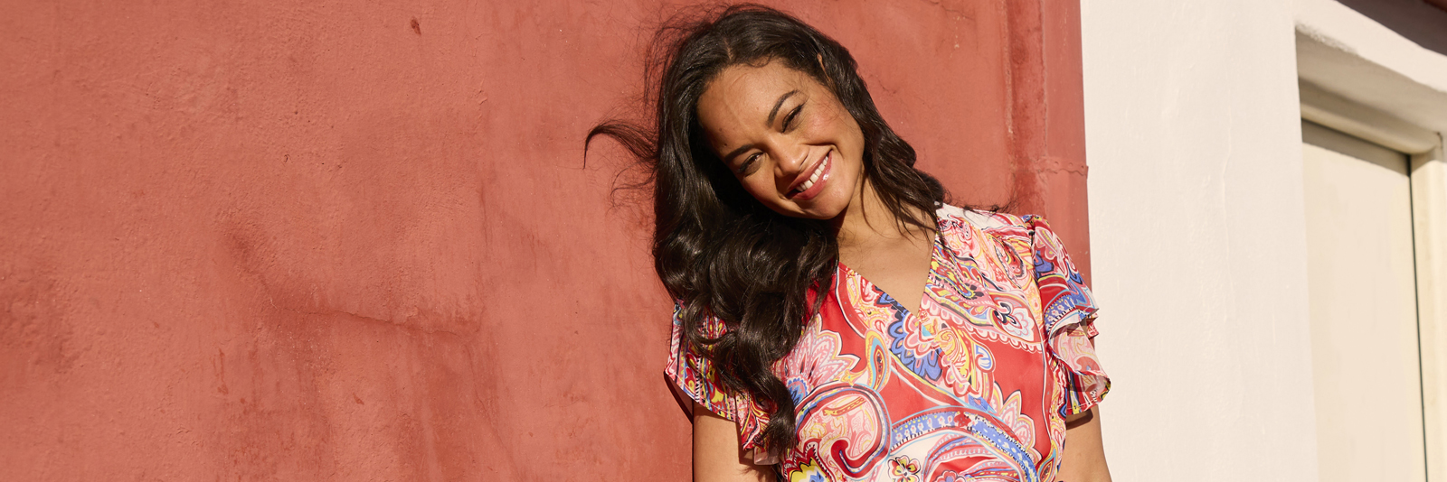 Frau lehnt mit einem sommerlichen Oberteil an einer Terracottafarbenen Wand