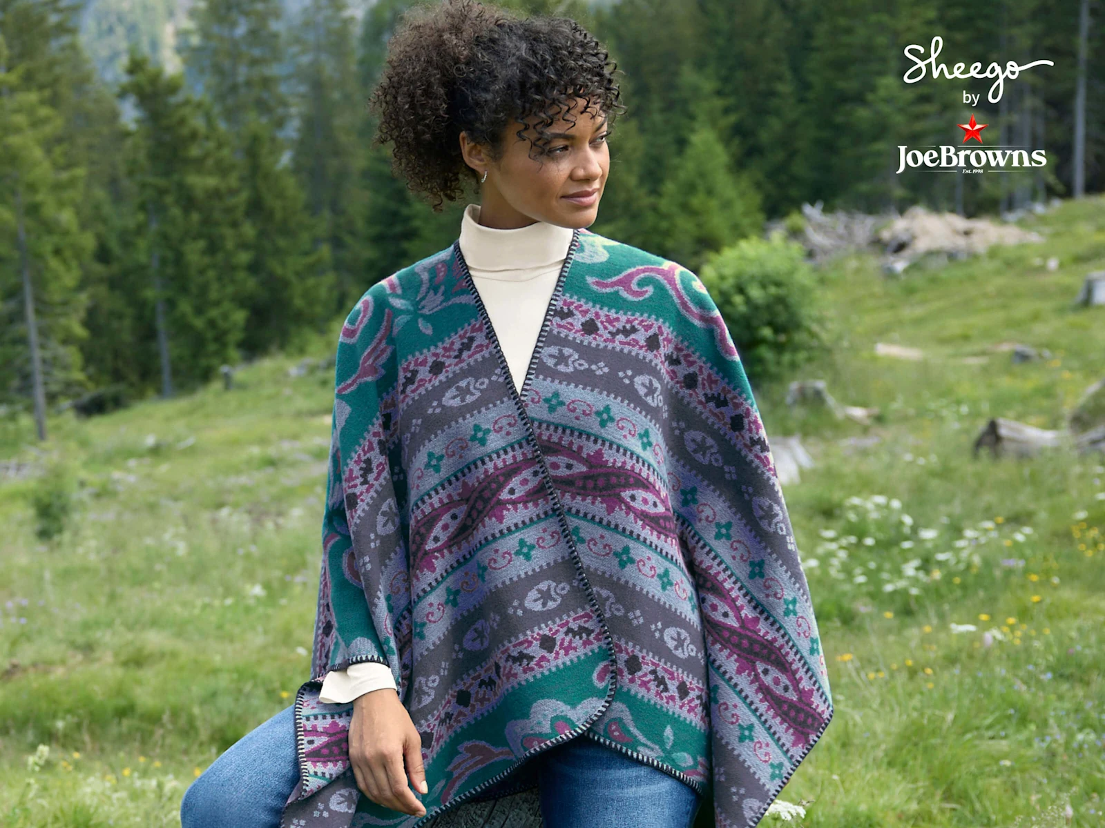 Ein Model trägt einen petrol-malve-gemustertn Poncho, ein champagnerfarbenes Rollkragenshirt und eine dark blue denim Bootcut-Jeans. Daneben ist das Sheego by Joe Browns Logo zu sehen und folgender Text: Warm ums Herz. Neues für den Winter