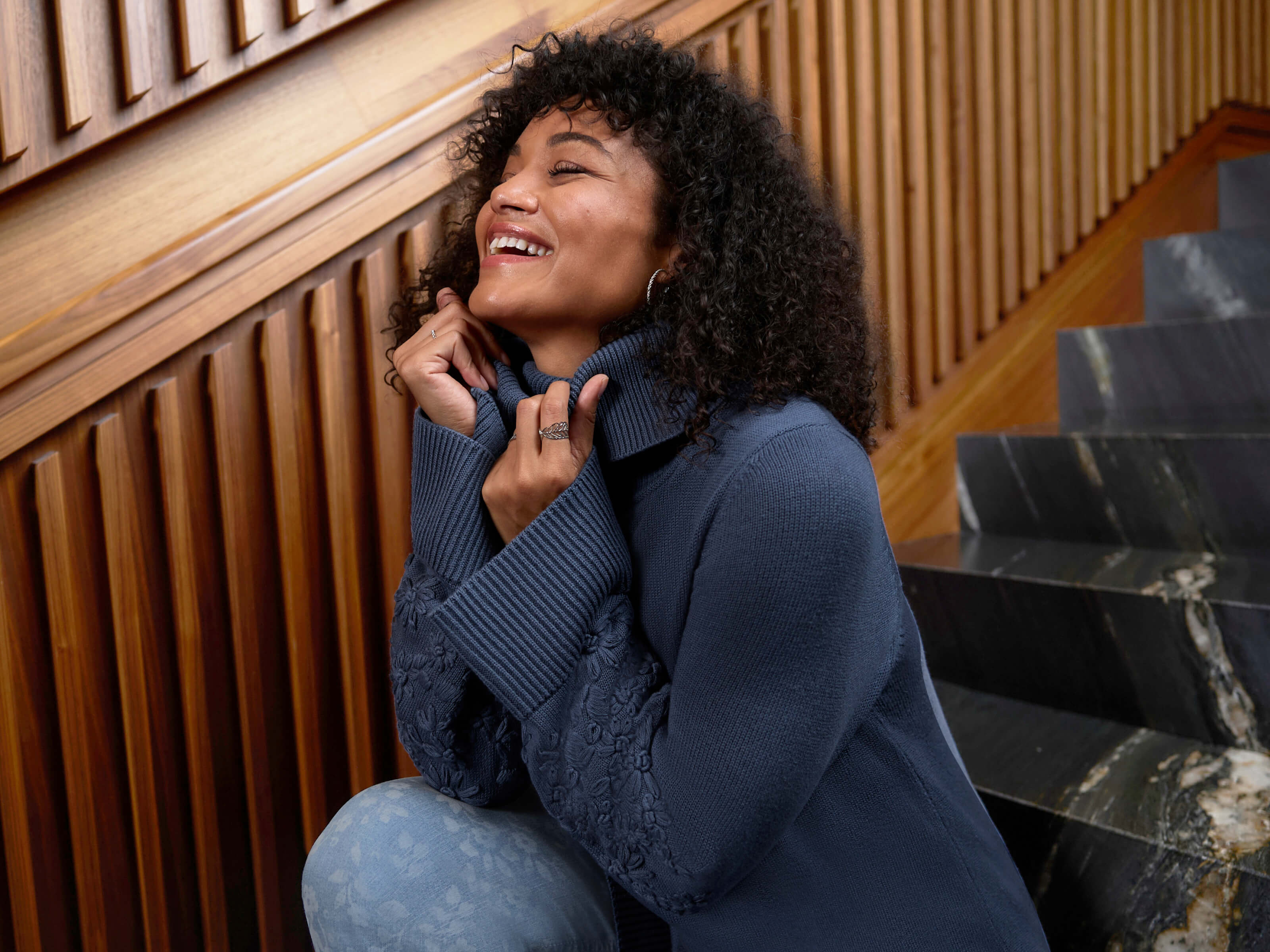 Ein Model trägt einen rauchblauen Rollkragenpullover mit Stickereien am Ärmel und eine blue bleached Bootcut-Jeans mit Laserprint-Blüten.