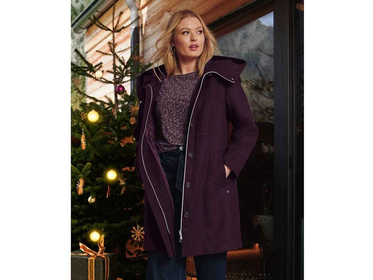 Ein Model in einer weihnachtlich geschmückten Umgebung. Sie trägt einen bordeauxfarbigen Kurzmantel
eine farblich passende Bluse und eine dunkelblaue Jeans.