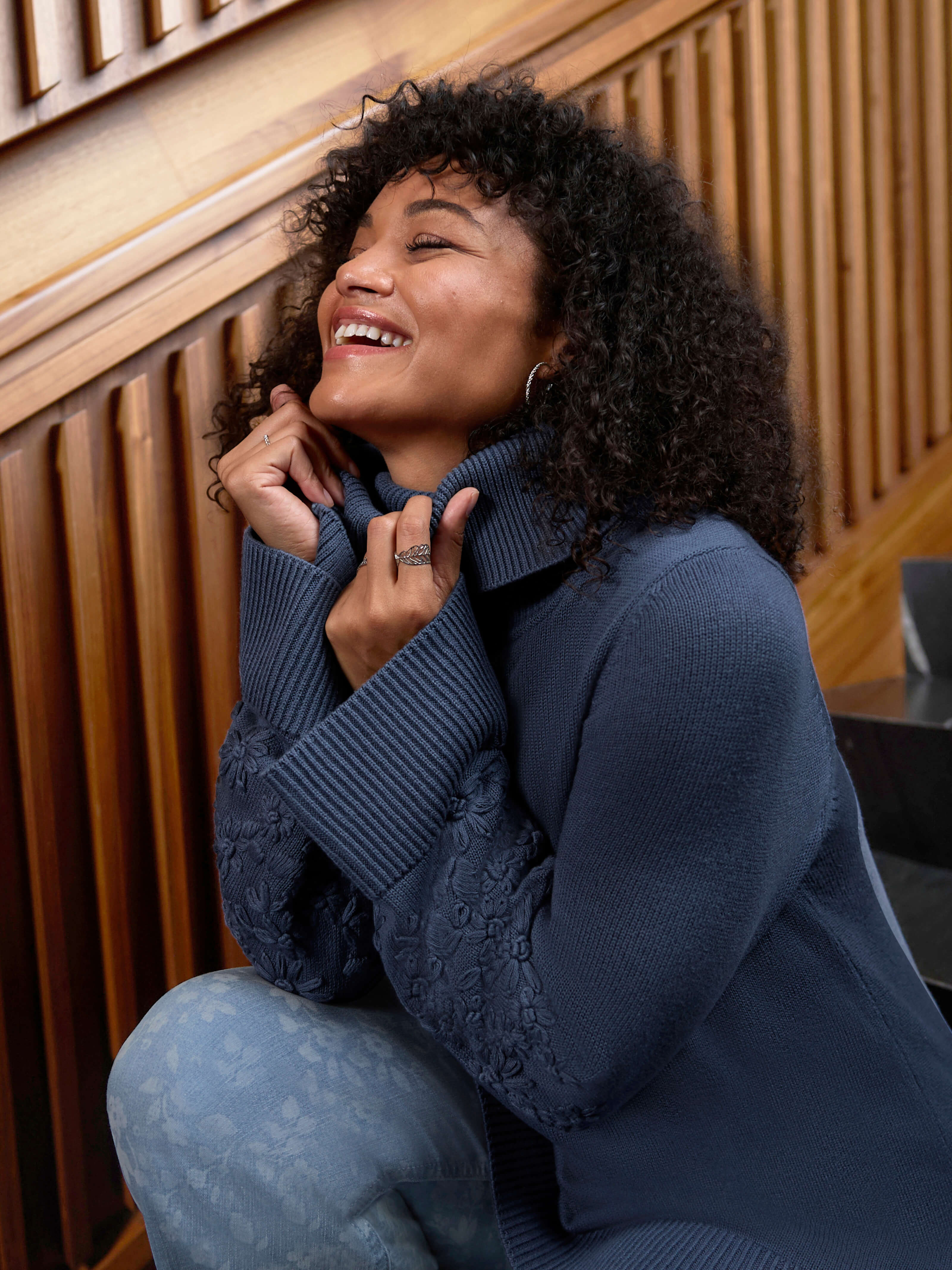 Ein Model trägt einen rauchblauen Rollkragenpullover mit Stickereien am Ärmel und eine blue bleached Bootcut-Jeans mit Laserprint-Blüten. Darunter folgender Text: Dark Mystic - Ein Hauch Geheimnis