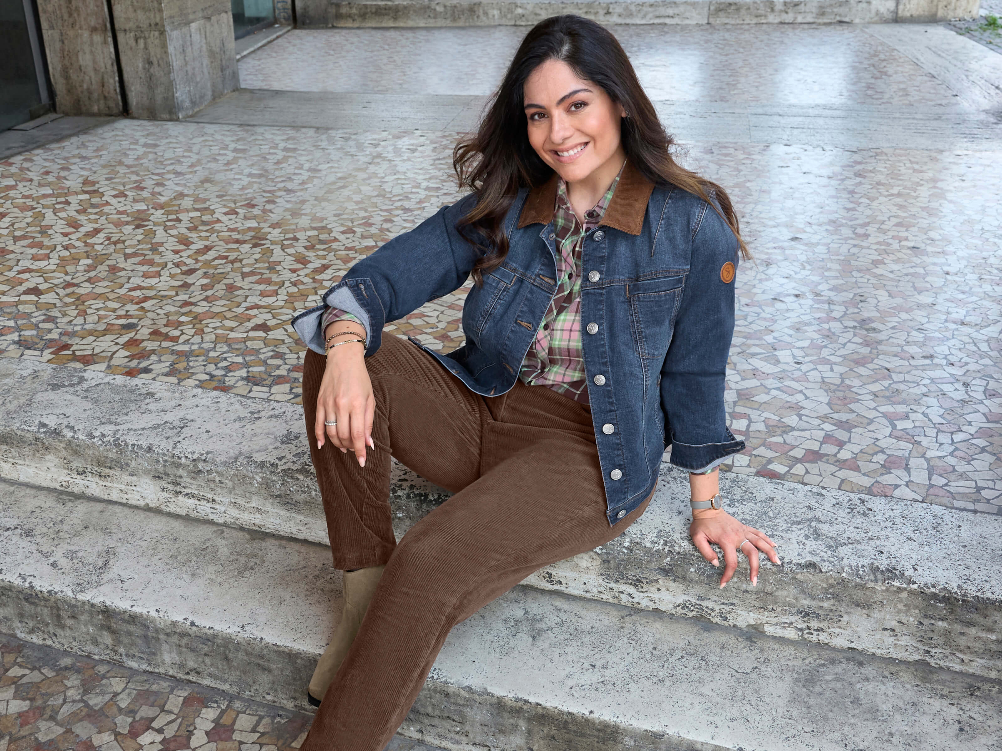 Ein Model trägt eine blue-stone-washed​ Jeansjacke​, eine schoko-hortensie-kariert​e Bluse und eine schoko​ farbene Cordhose