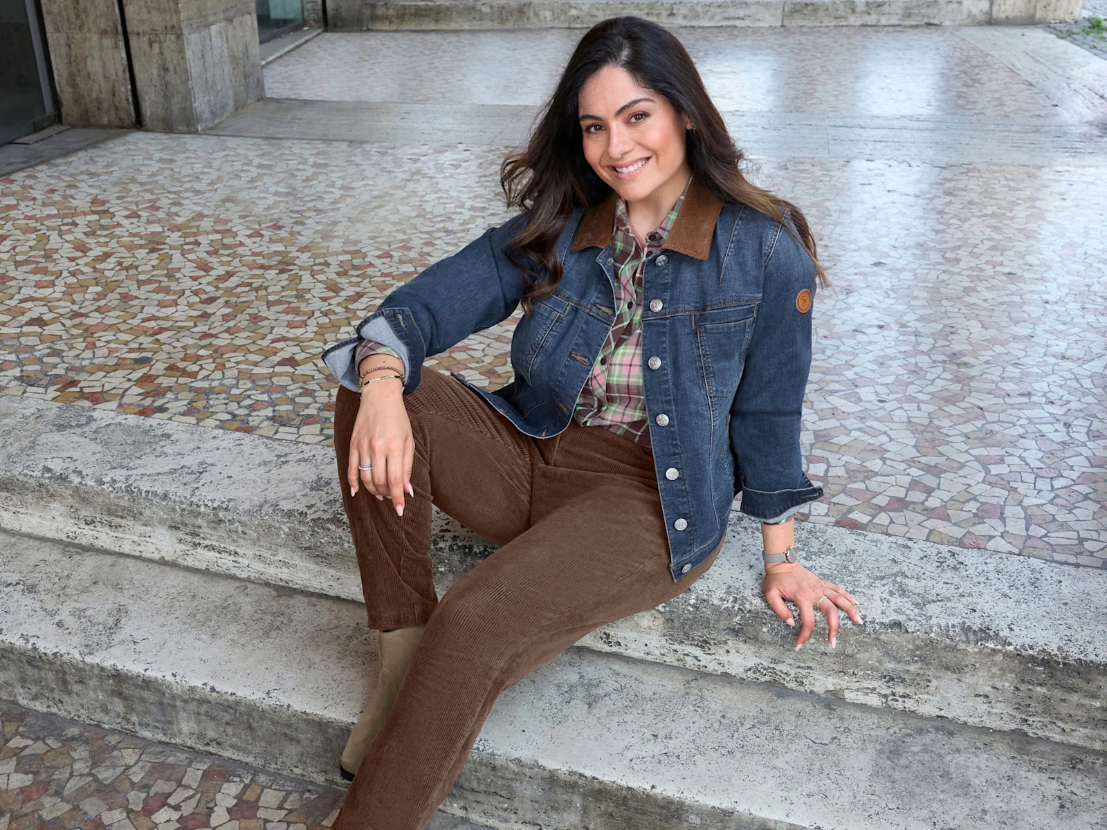Ein Model trägt eine blue-stone-washed Jeansjacke, eine schoko-hortensie-karierte Bluse und eine schoko farbene Cordhose