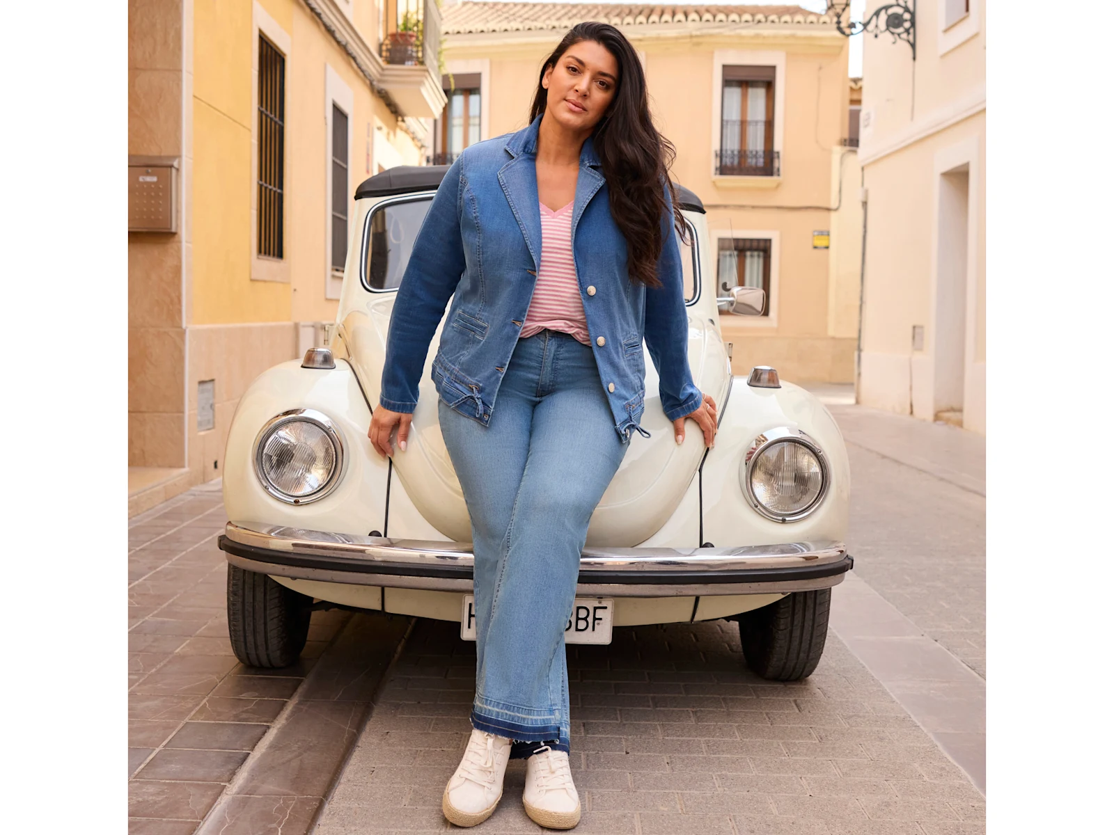 Ein Model in einer City-Umgebung, sie lehnt an einem Auto und trägt einen blue bleached Jeansblazer, ein ecru-rosa gestreiftes Shirt und eine blue-bleached Bootcut Jeans.