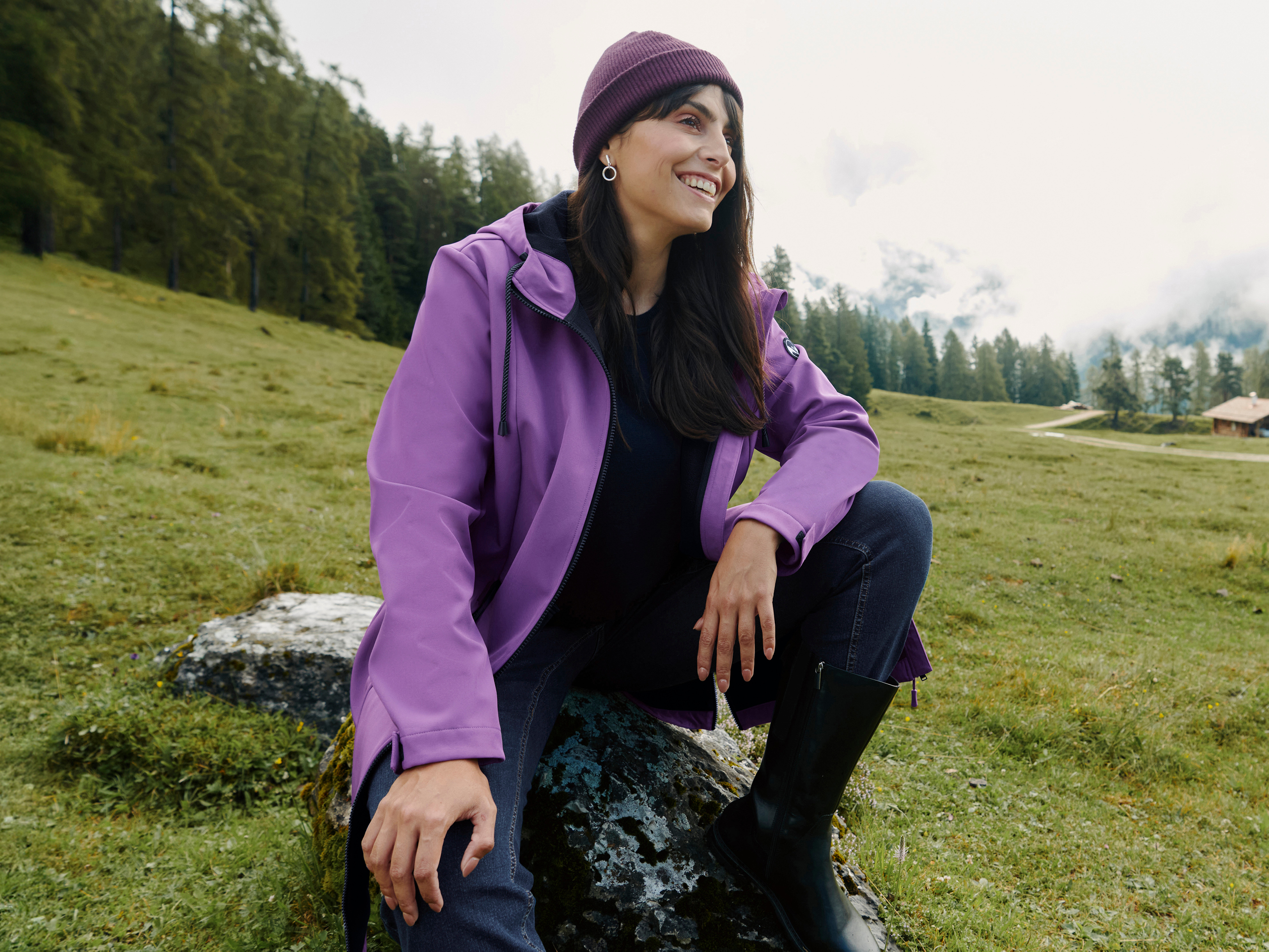 Ein Model sitzt auf einem Stein in einer grünen Wiese, trägt eine lila Softshelljacke, Mütze, Jeans und schwarze Stiefel.