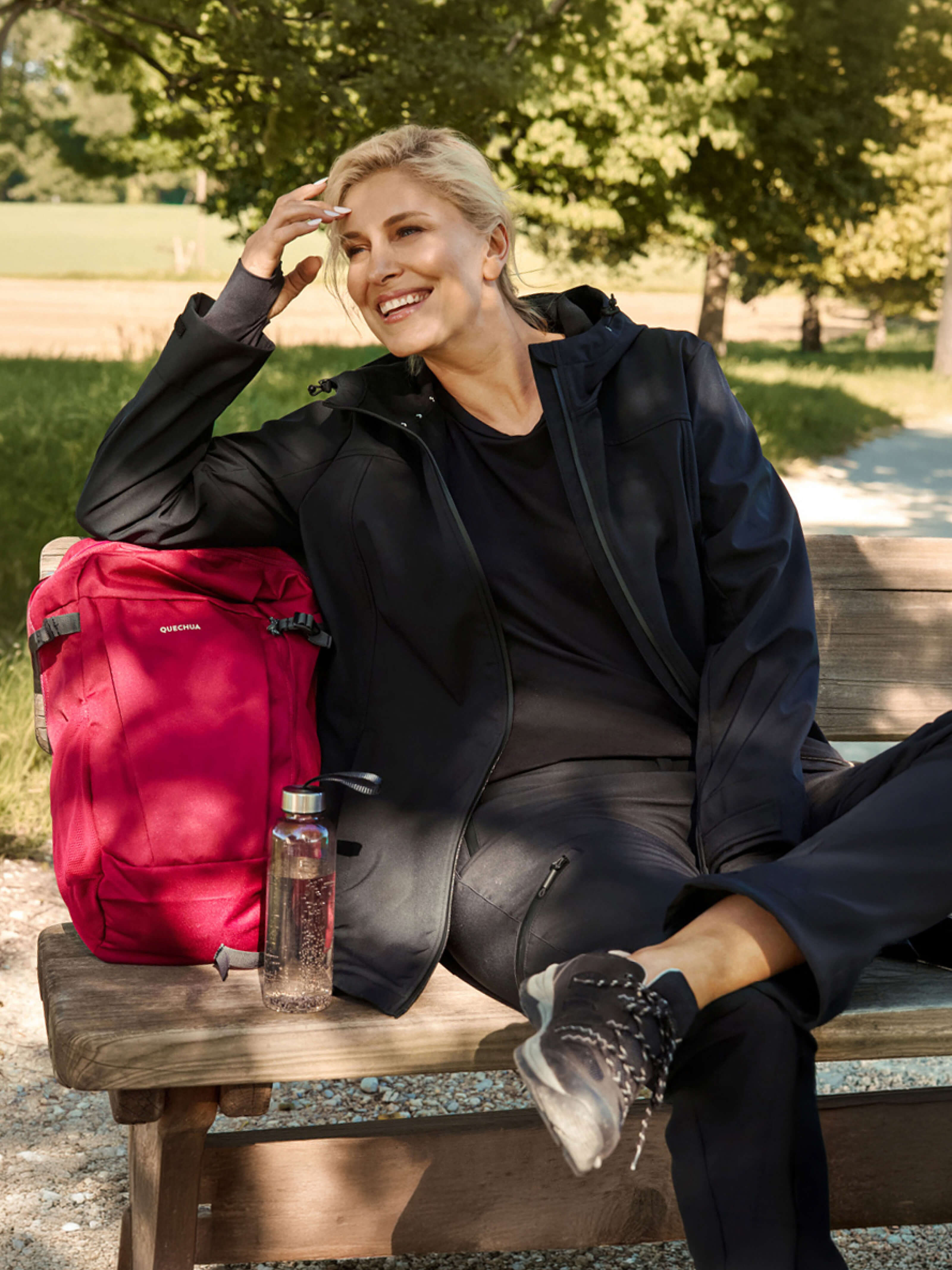 Ein Model im Wander-Outfit sitzt auf einer Bank, sie trägt eine schwarze Softshelljacke, dazu passende Hose und Wanderschuhe. Daneben folgender Text. An die frische Luft in funktionalen Sport-Styles
