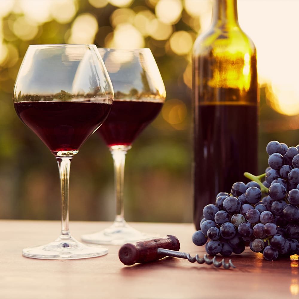 Two glasses of red wine next to the bottle, a bunch of grapes and a cork screw on a table