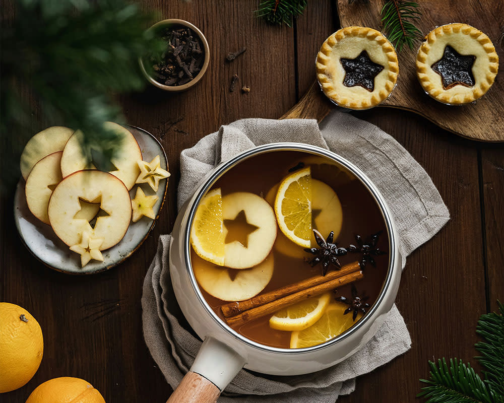 a saucepan of cooking mulled wine with spices and fruit next to mince pies