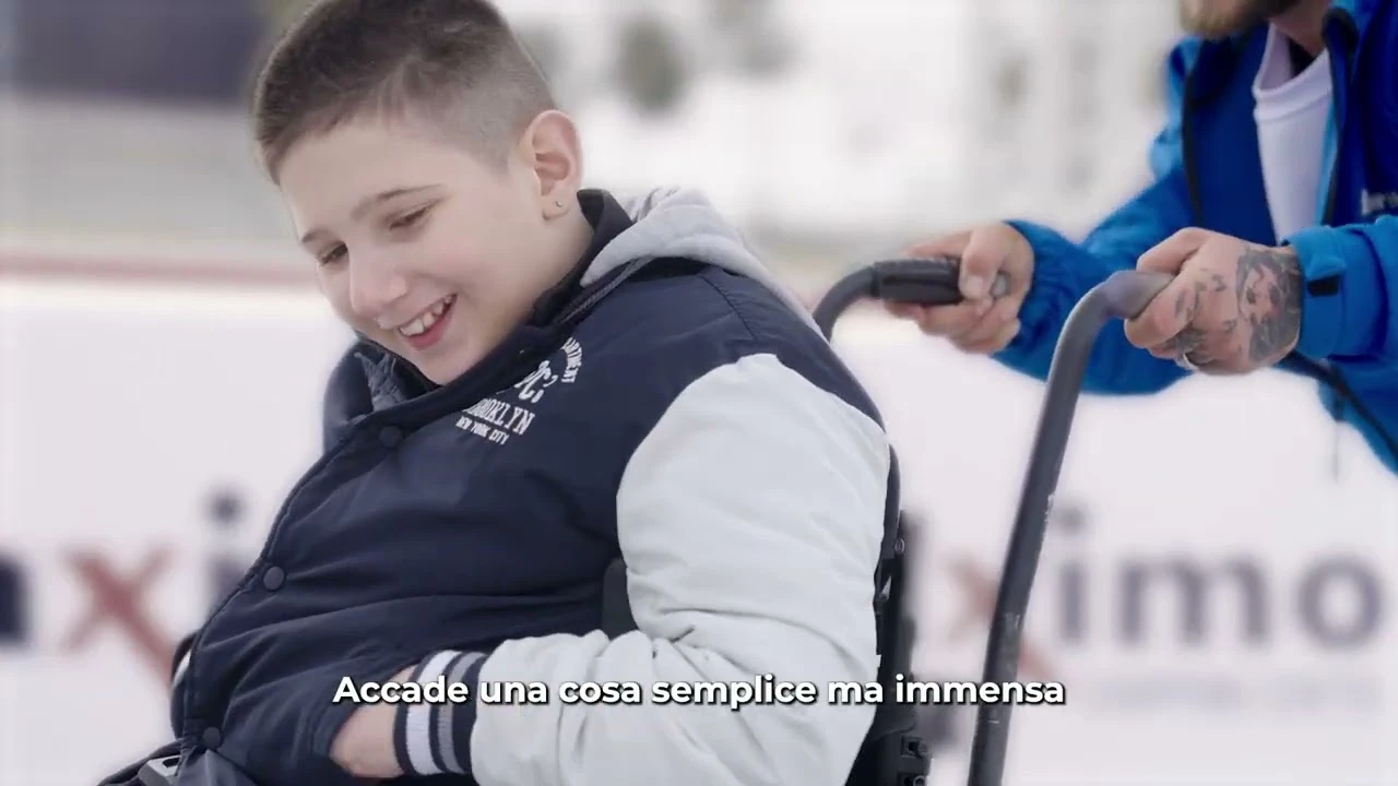 Bambino seduto su una sedia a rotelle su una pista di ghiaccio, spinto da un’altra persona.