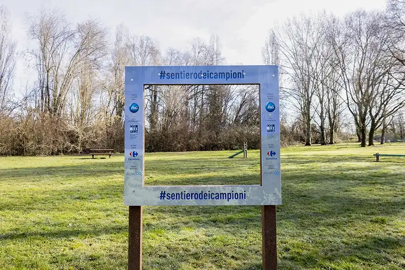 Un’immagine di un parco con un prato verde e una cornice che riporta i loghi di P&G, Corepla e Carrefour e il nome del progetto “Il Sentiero dei Campioni”.