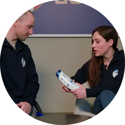 Due persone in uniformi scure sono sedute in un ambiente interno, e una di loro mostra una bottiglia di prodotto all’altra