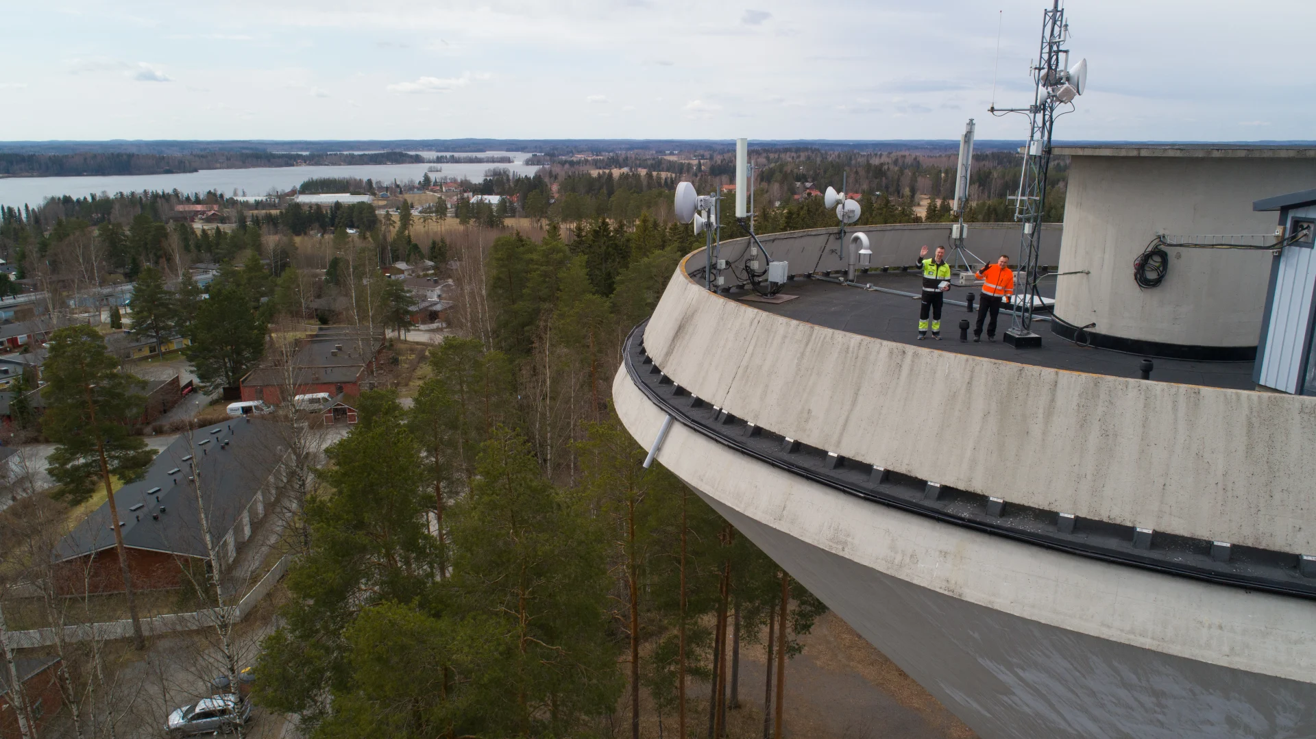 Instan työntekijä ja asiakas ilmasta kuvattuna Lempäälän vesitornin huipulla, tornista osa rajautuu pois ja vasemmalta näkyy taustan maisemaa. 