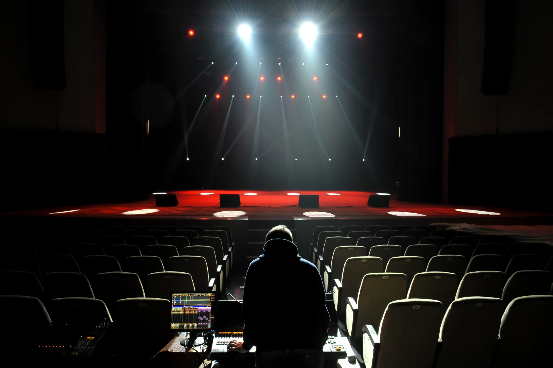 The stage of the theater is photographed from the front. In the foreground of the picture is a theater technician at the control table with his back to the camera.