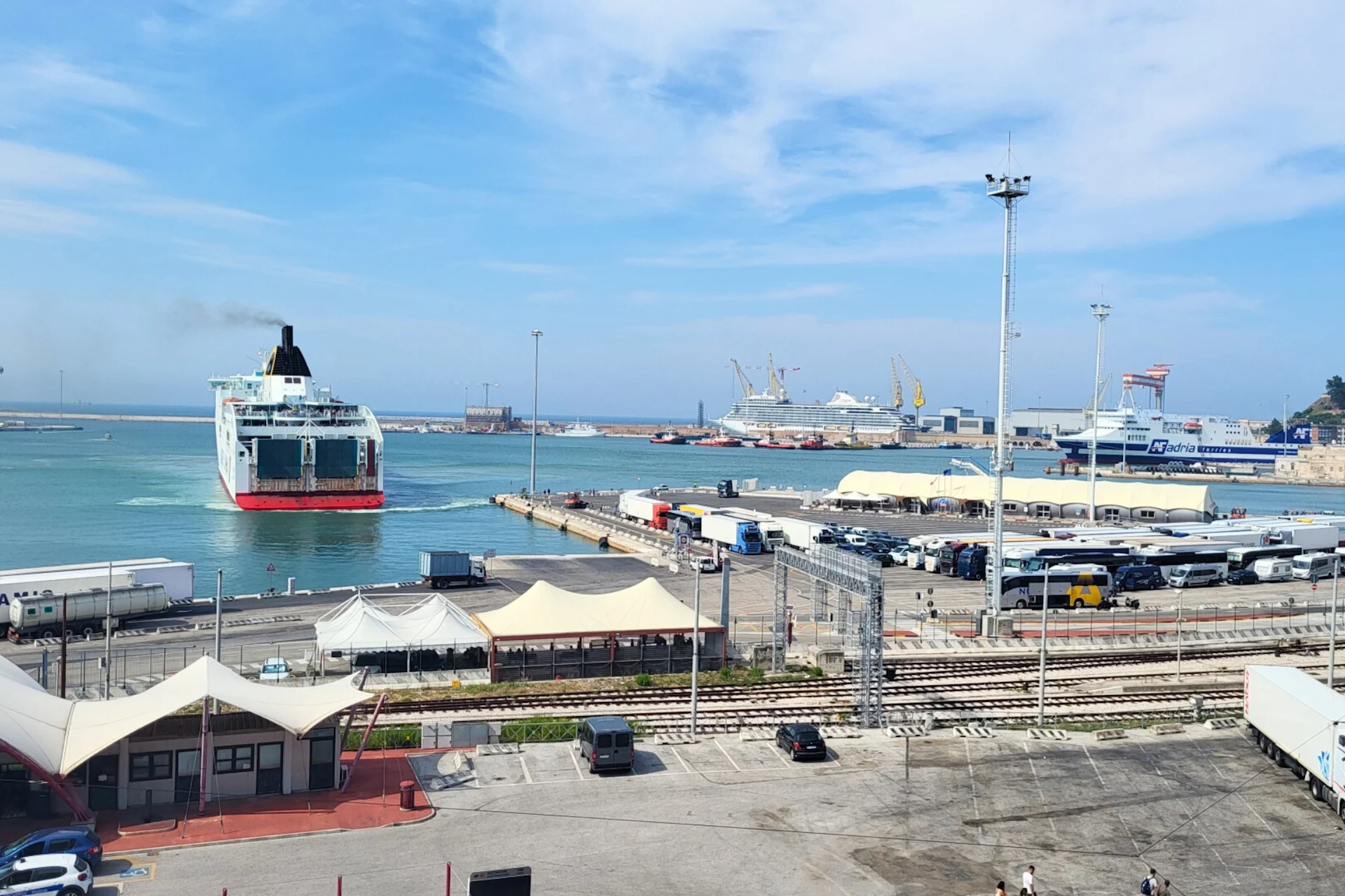 A passenger vessel departing a port area surrounded by industrial harbor structures and logistics facilities.
