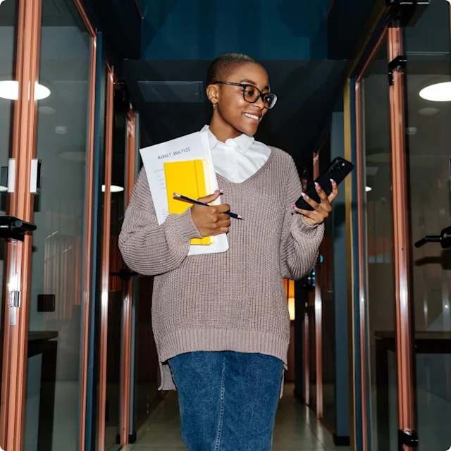 Woman looking at phone walking in an office environment with notes in her other arm.