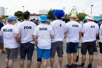 6 men are lined up with their backs to the camera. They are all wearing white t-shirts with words on the back spelling out BLUE FOR BOB 8 FOR 43. 5 of them are wearing hats, and one is wearing a bright blue curly wig. 