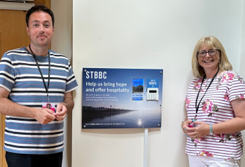 Rev David Mayne and Rev Jo Hellenbrand, pastors at Shoebury and Thorpe Bay Baptist Church standing next to a wall mounted point of donation that uses Give A Little's DIY approach
