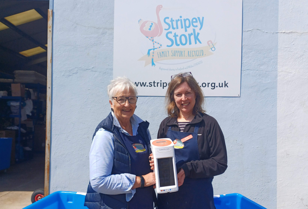 Photo of two members of staff at Stripey Stork standing outside the charity's sign and holding a CollecTin More contactless donation box that uses Give A Little software 