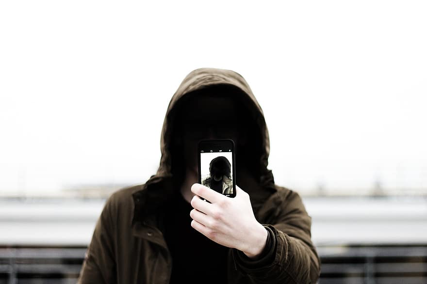 Zakapturzona osoba robiąca sobie selfie. Nie widać twarzy, czarny kaptur, smartfon z włączonym aparatem.