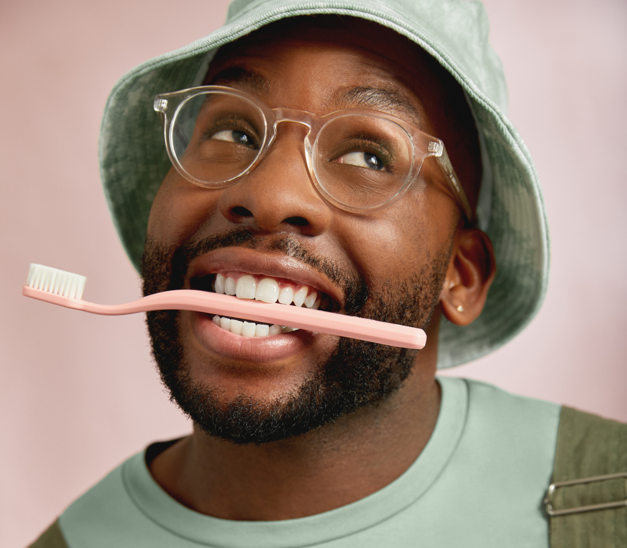 Man in hat smiling and looking upward with toothbrush handle being held by his teeth