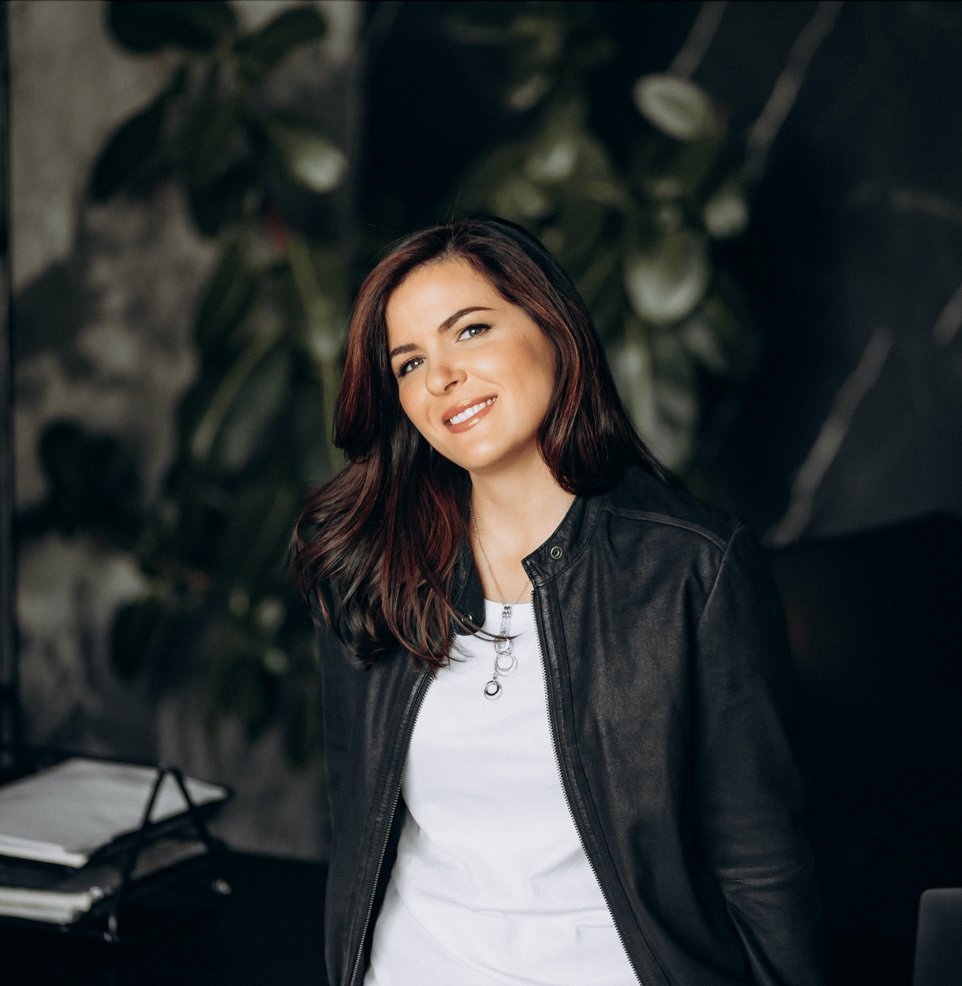 The picture reflects the author's portrait - the white smiling woman, around 30 years old. The woman is wearing a white T-shirt and a black leather jacket. She has long brown hair with red-colored stripes and natural makeup. The woman is standing next to the office table with some working papers on it. The background of the portrait is dark green and grey.