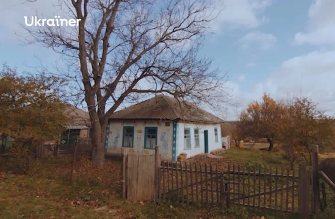 Типовий український сільський будинок початку ХХ століття з високим деревом поруч з будинком. Перед будинком старий тендітний паркан. Фотографія зроблена в осінню пору.