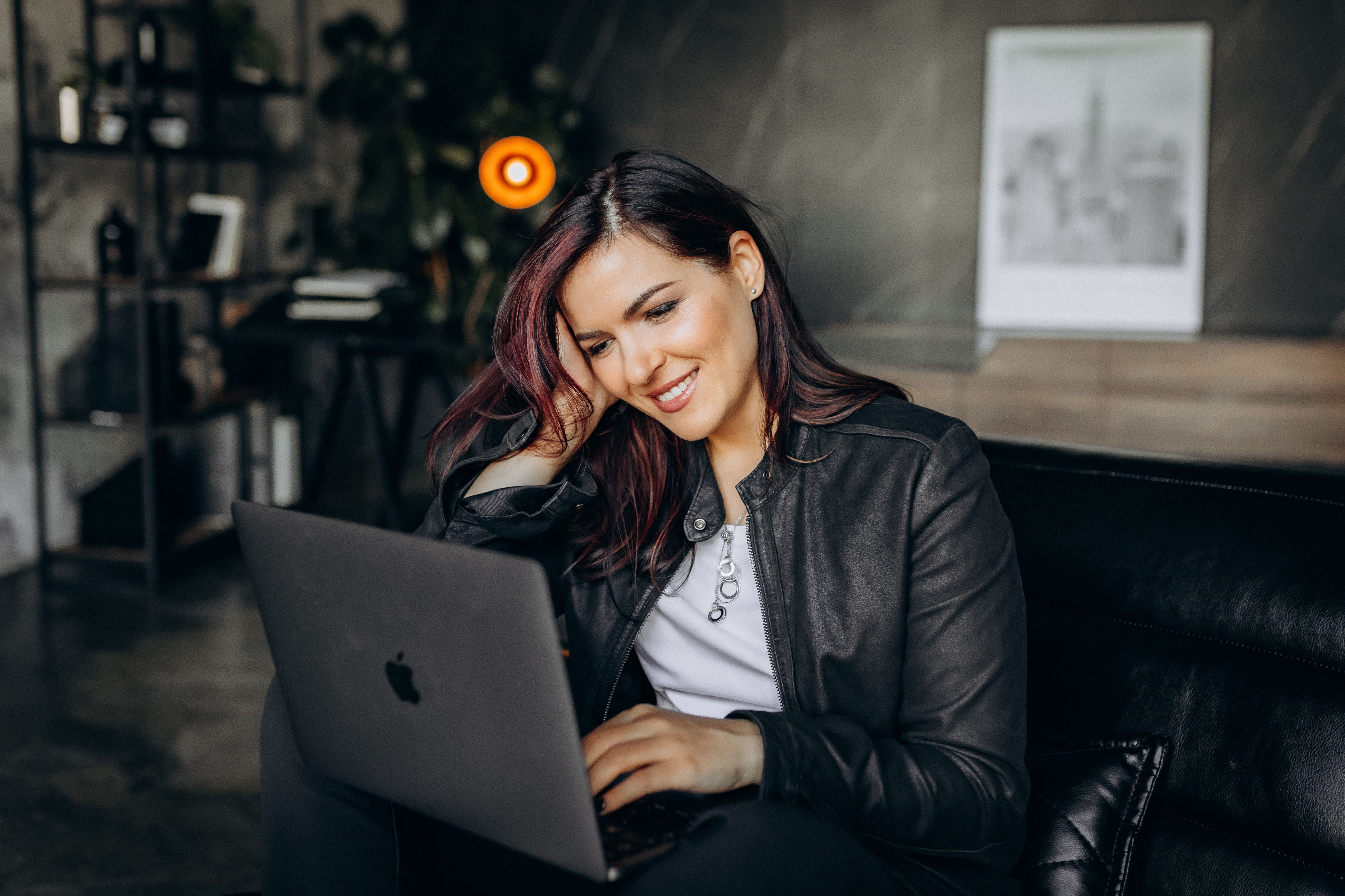 The photo reflects the smiling author sitting in the black leather chair and looking at her laptop. She holds her head with her right hand while her left hand is on the keyboard of the laptop. In the background, on the left, there are carcass-style shelves with books, flowers, and accessories. Behind the author, there is a table with a big picture of some megapolis panorama.