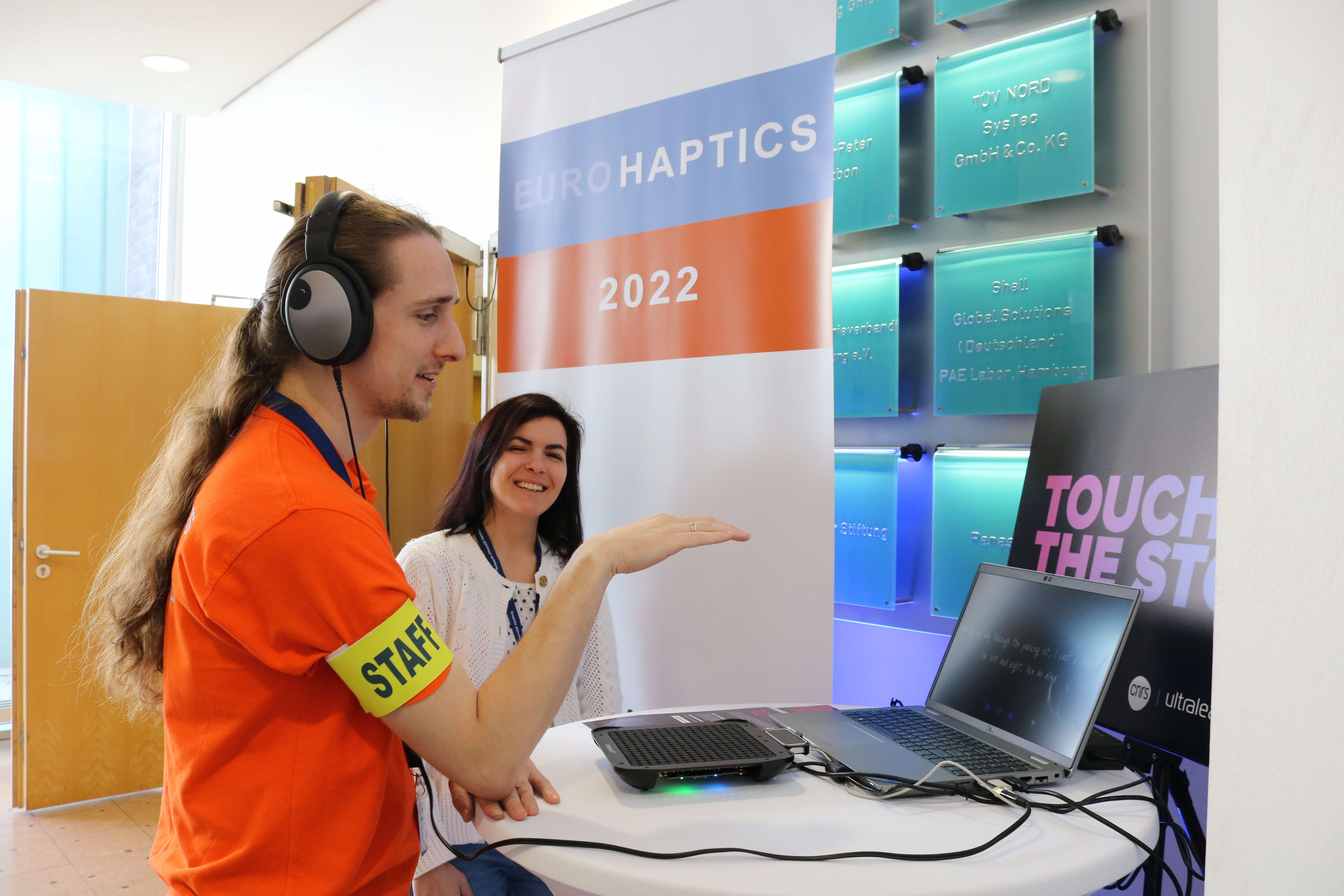 The author is demonstrating the solution at EuroHaptics 2022 conference. The picture shows the man experiencing “The Touching Story“ and the author standing next to him. The man is wearing acoustic headphones, looks at the screen displaying text and holding his hand over the haptic device.