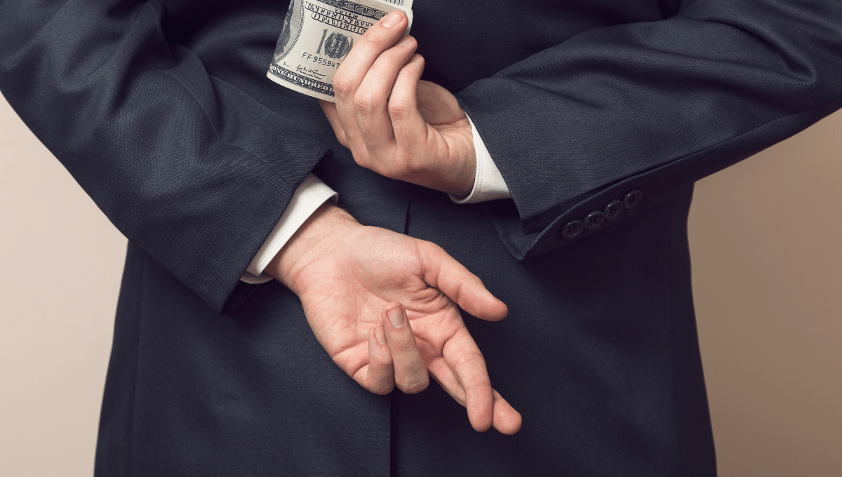 white employee in a black suit with crossed fingers holding a wad of cash behind back