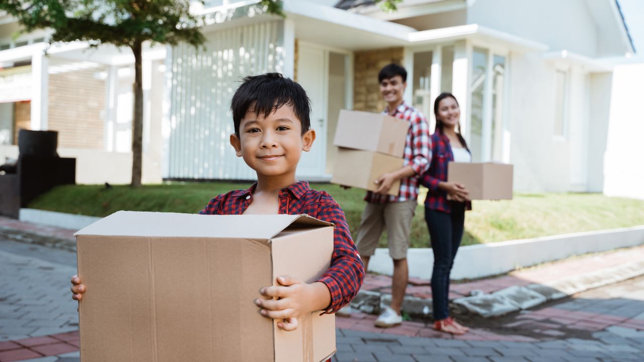 A small boy with dark skin and hair stands in the driveway of a house holding a cardboard moving box, with his parents in the background also holding boxes.